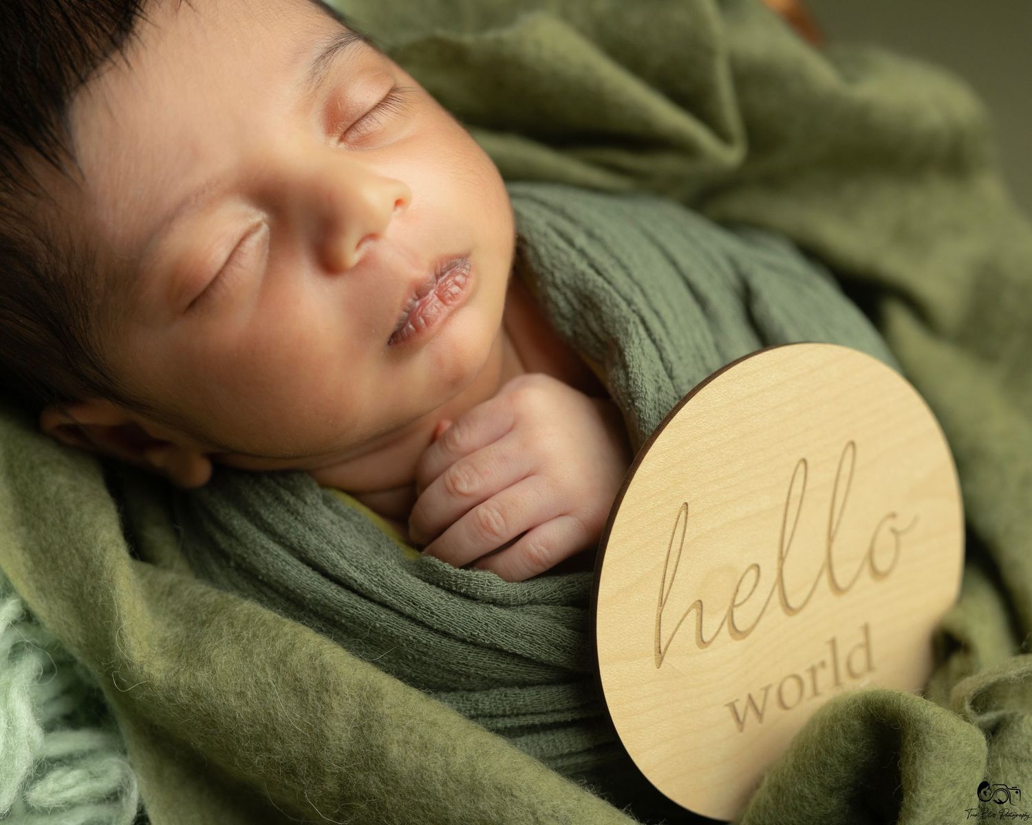 Peaceful newborn baby sleeping wrapped in green blanket holding a wooden hello world disc milestone sign.