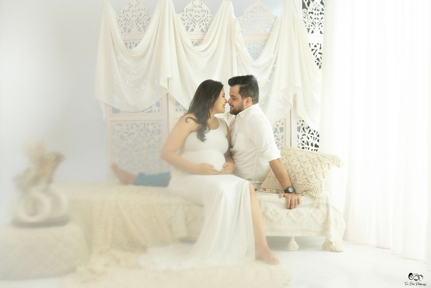 Couple sharing a tender moment in a bright white studio, pregnant woman in flowing white gown with partner during maternity photoshoot.