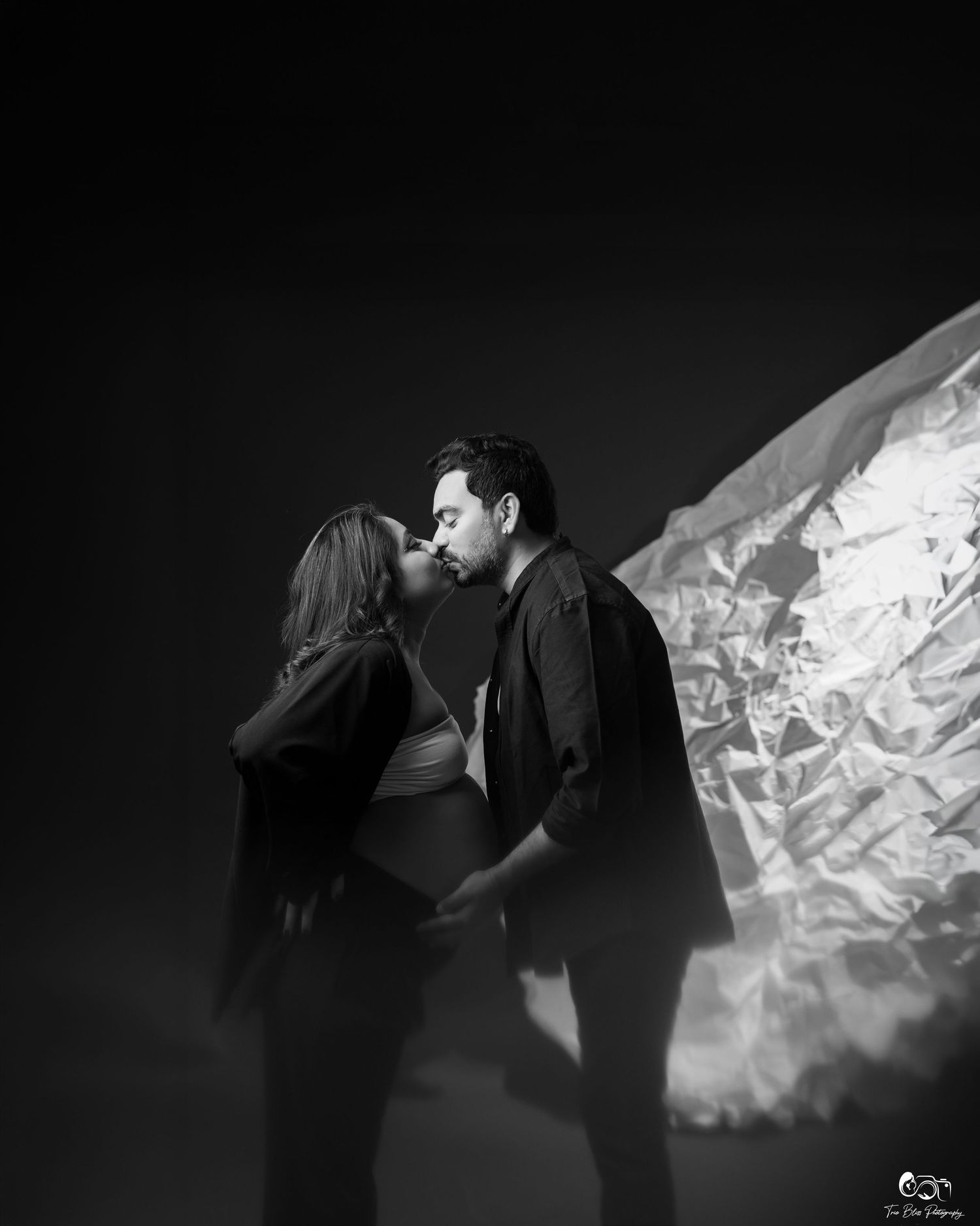 Black and white photo of a couple kissing during a maternity shoot with dramatic light and shadow backdrop.