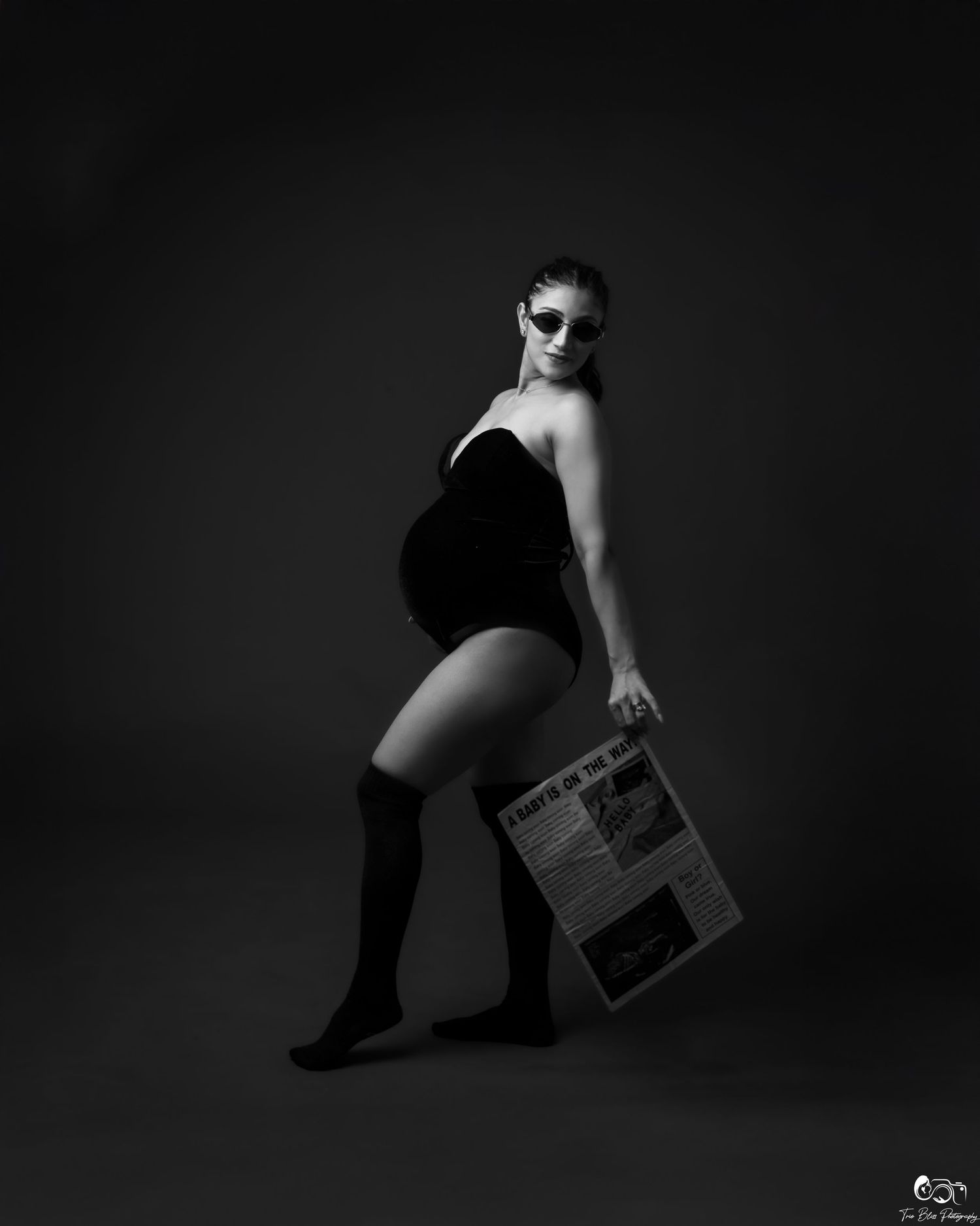 Pregnant woman in black swimsuit and sunglasses poses dramatically holding a newspaper in a black and white studio photo.