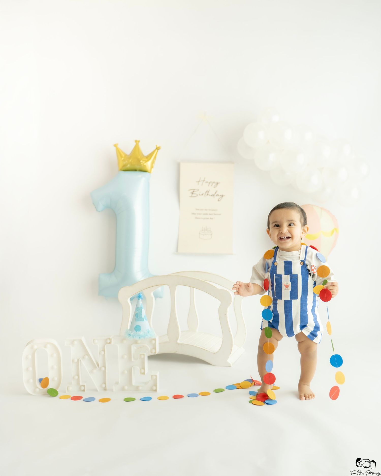 Baby's first birthday photo with number one balloon, crown, ONE letters, colorful garland, and white decor backdrop.