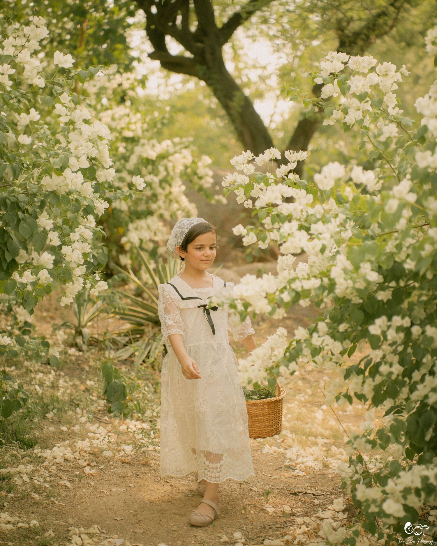 A young girl in a white lace dress holds a basket among blooming white flowers in a dreamy garden setting.