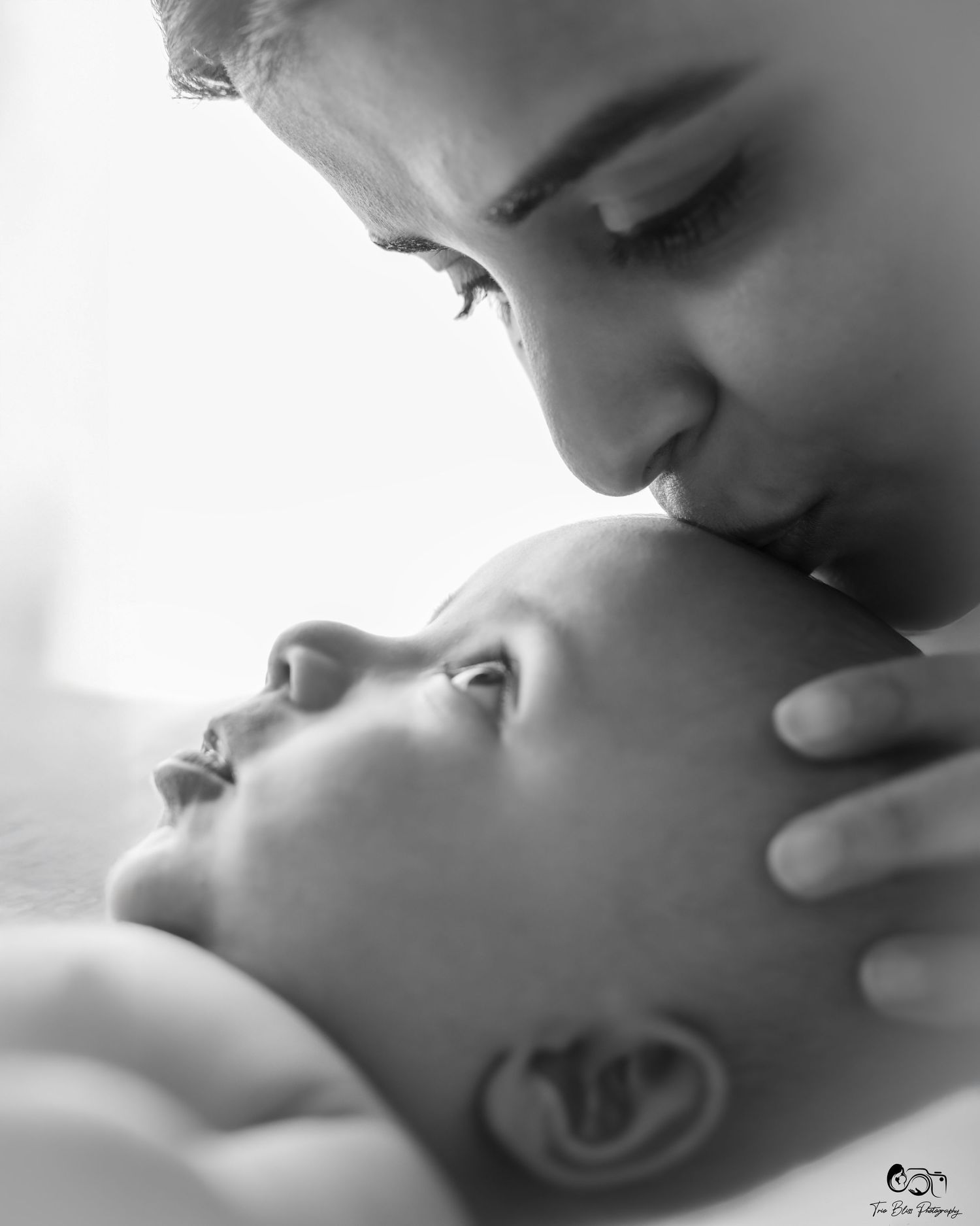 Black and white photo of a parent tenderly kissing a newborn baby on the forehead in an intimate moment.