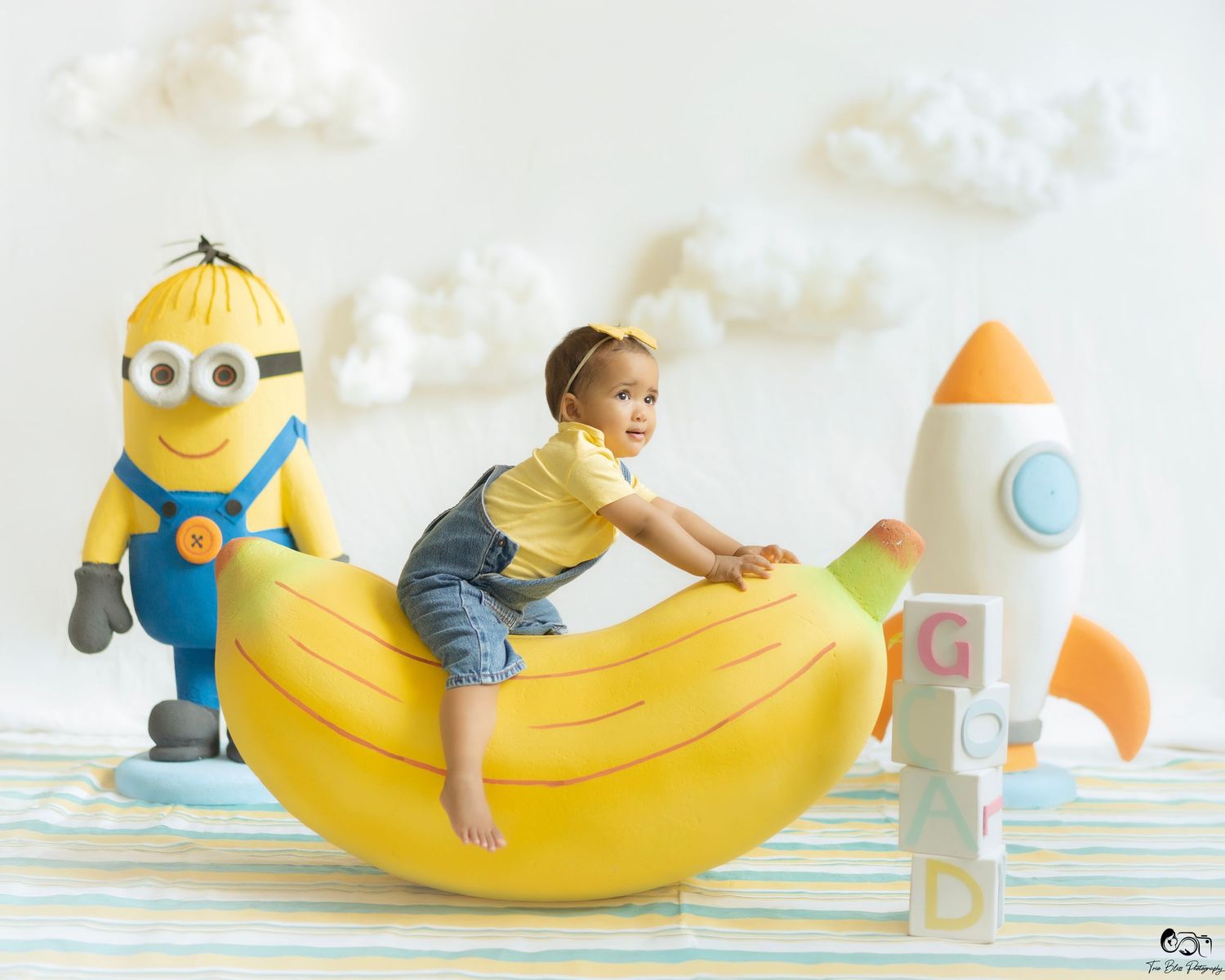 A smiling baby sits on a giant banana toy beside a Minion plush and a toy rocket with alphabet blocks.