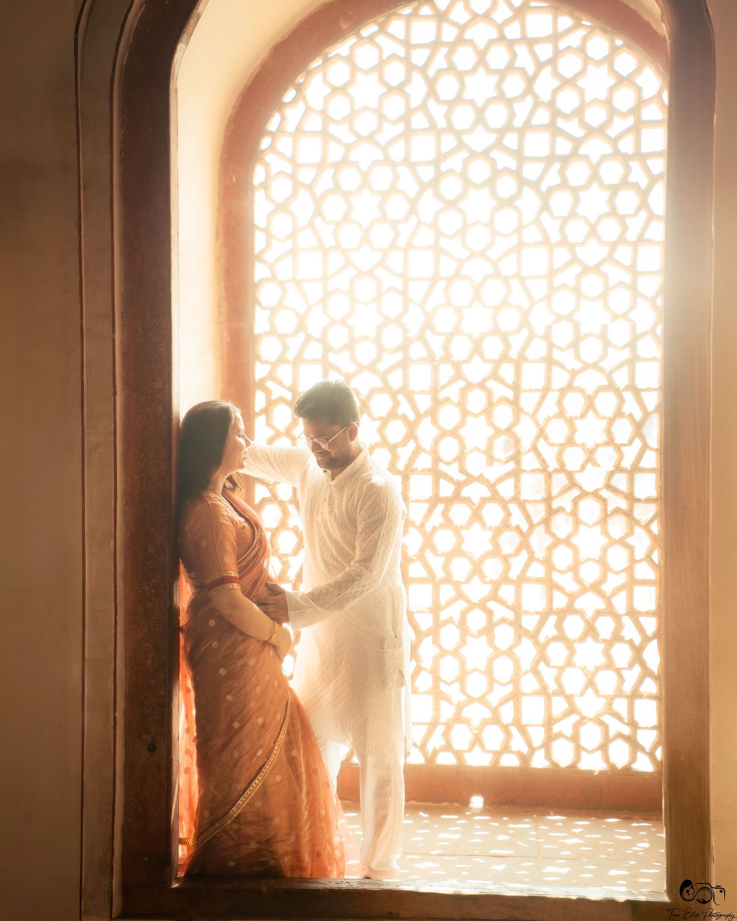 Couple in traditional Indian attire standing in a glowing arched Mughal window, romantic pre-wedding shoot.