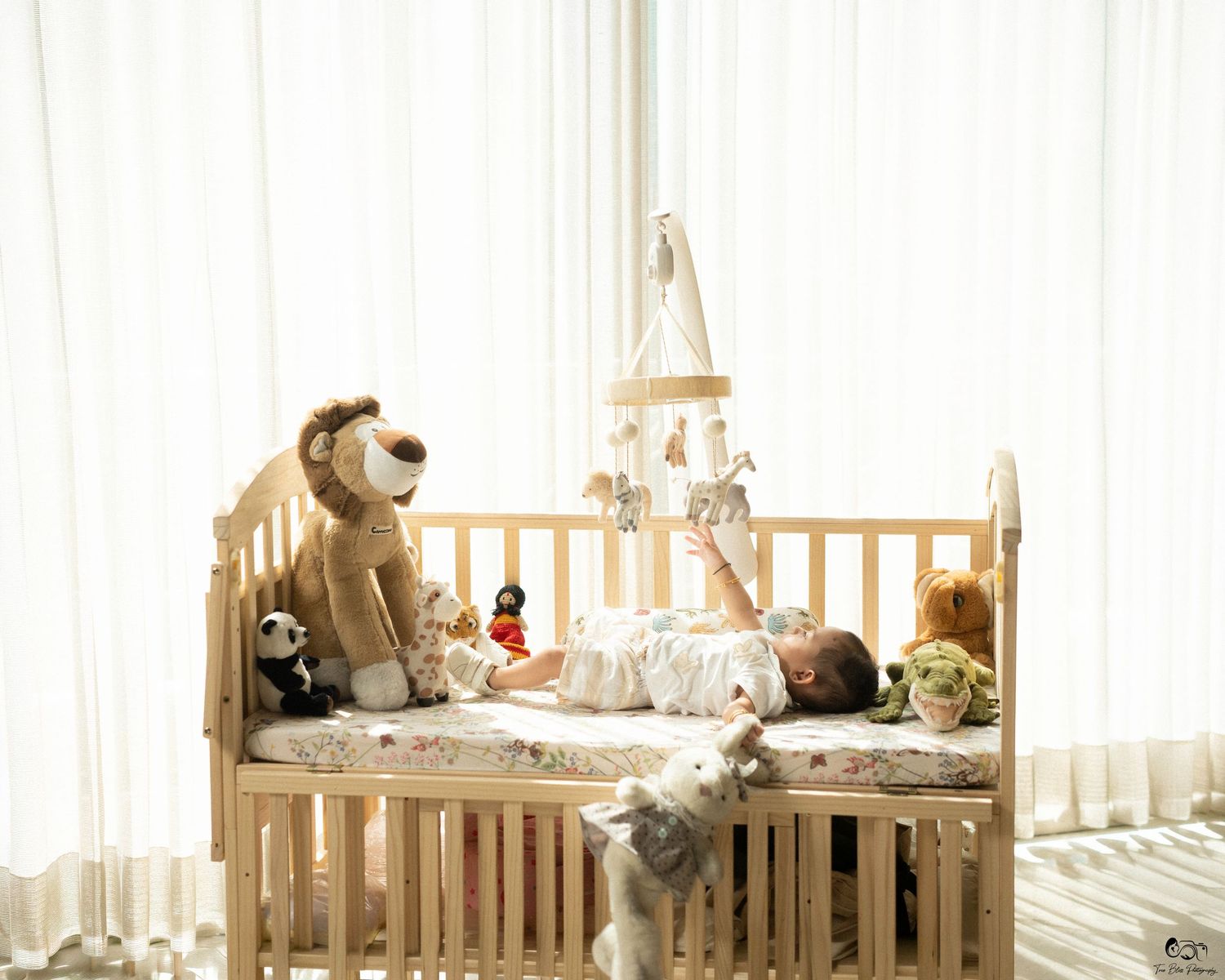 Baby lying in wooden crib surrounded by stuffed animals with a mobile hanging above in a bright nursery.