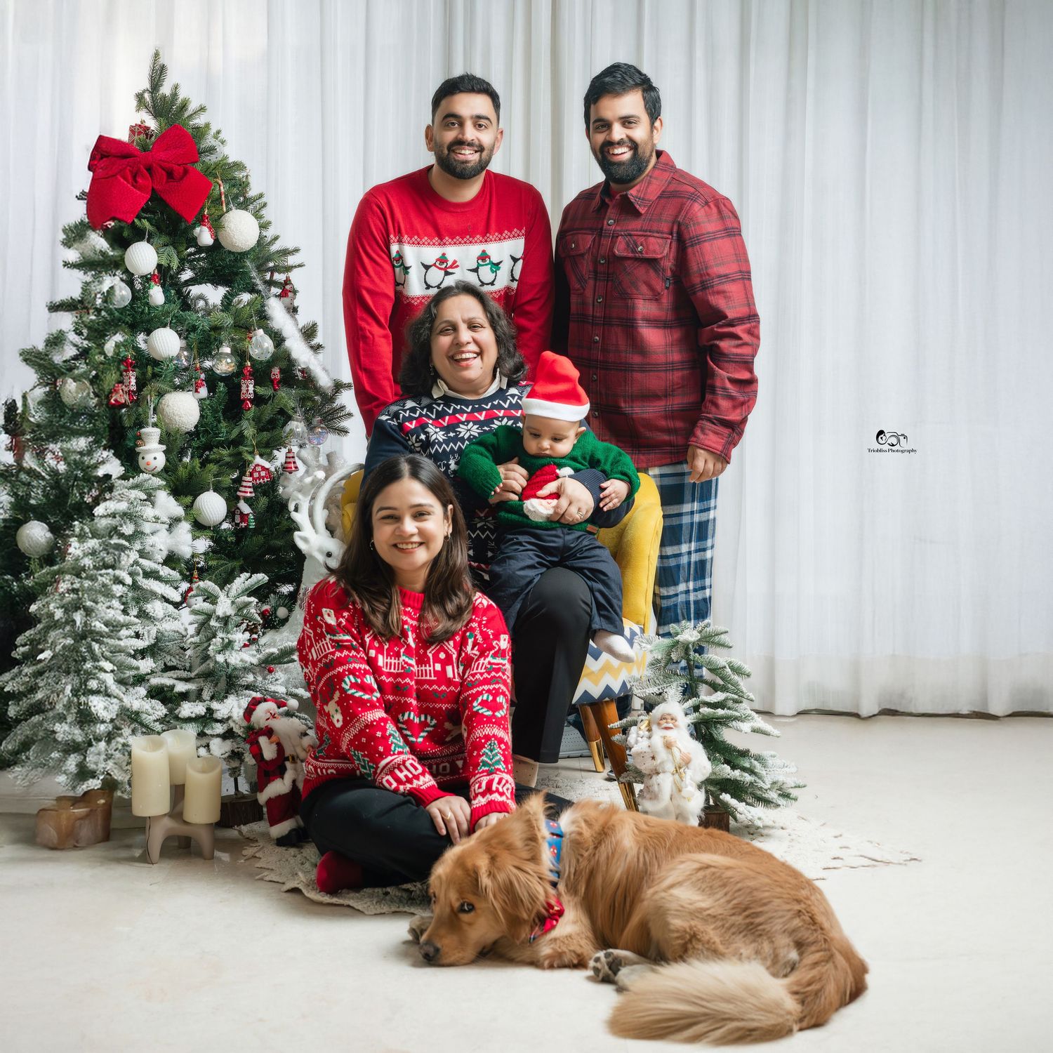 Family in Christmas sweaters posing with decorated tree, baby, and golden retriever dog in festive holiday photo.