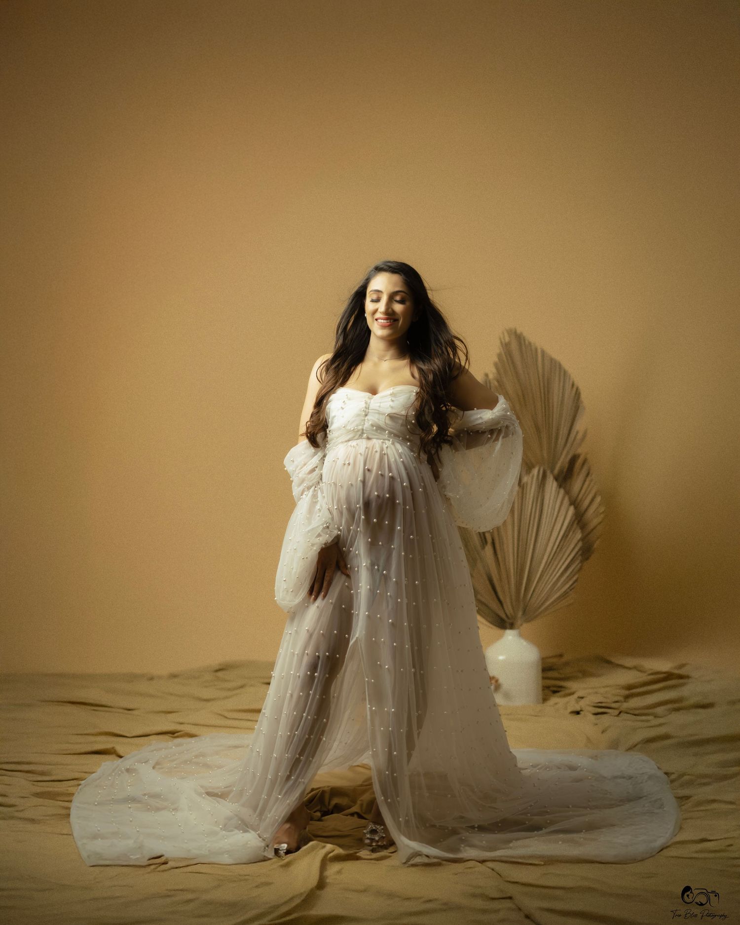 Pregnant woman in white tulle maternity gown posing gracefully in a warm beige studio setting.