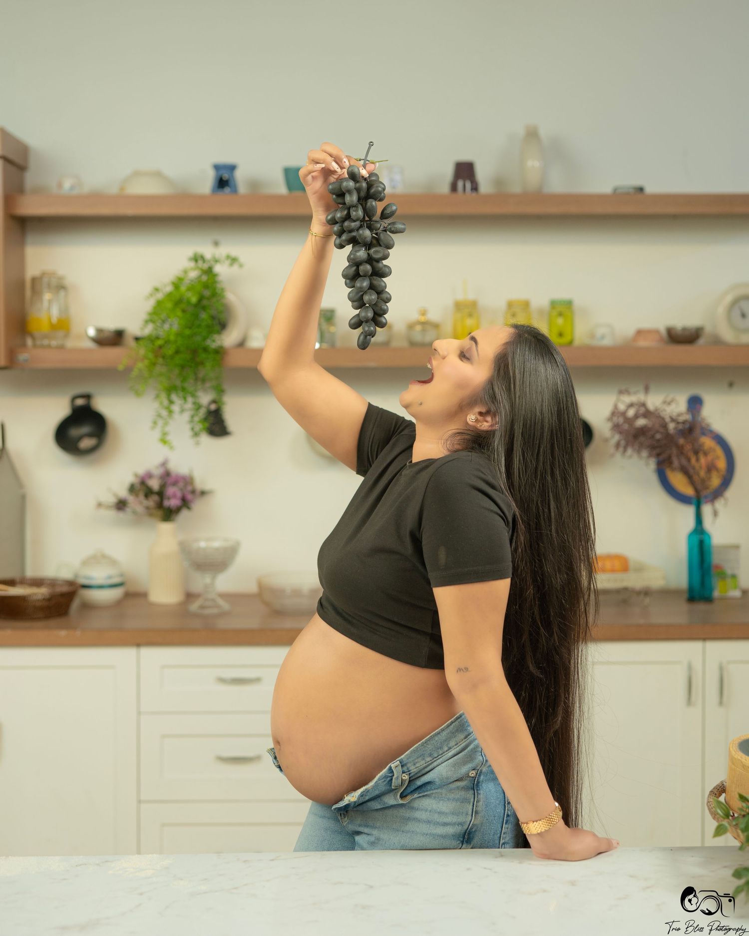Pregnant woman in black crop top and jeans holds grapes above her mouth in a bright modern kitchen.