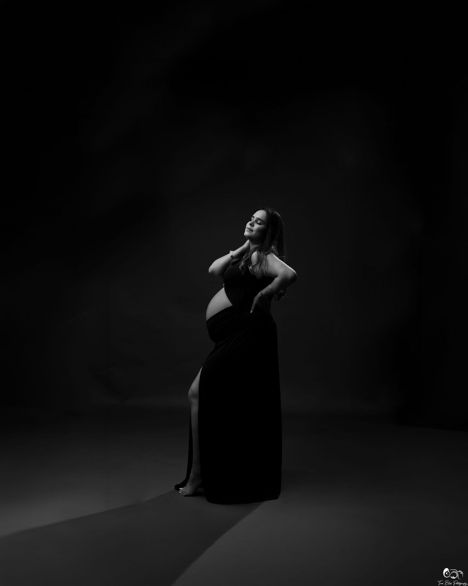 Black and white maternity photo of a pregnant woman in a dark gown posing gracefully in dramatic studio lighting.