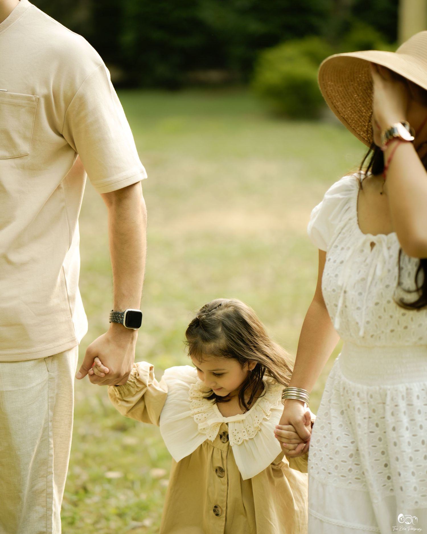 A young girl holds hands with her parents during an outdoor family portrait in a lush green park setting.