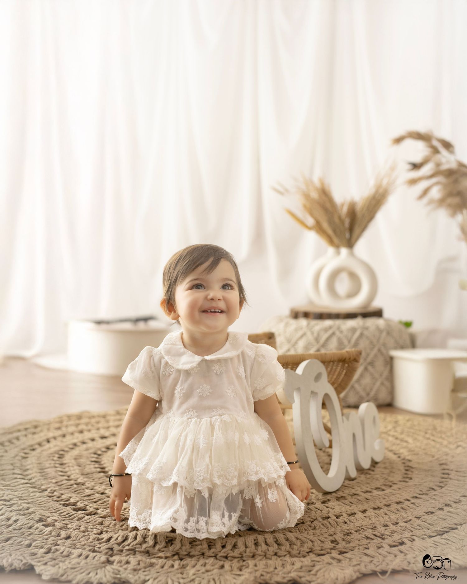 Smiling baby girl in white lace dress sits on jute rug beside wooden One sign in bright boho studio setting.