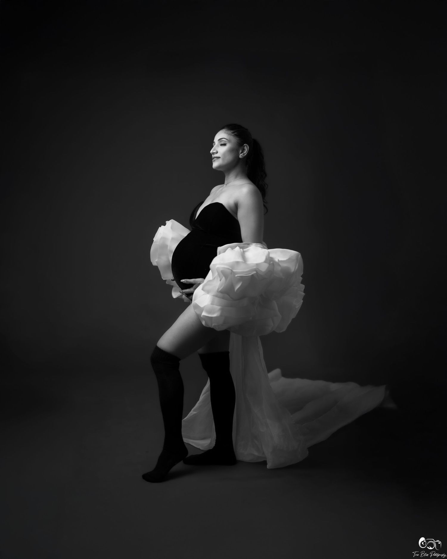 Black and white maternity photo of pregnant woman in strapless top, ruffled skirt, and knee-high socks posing gracefully.