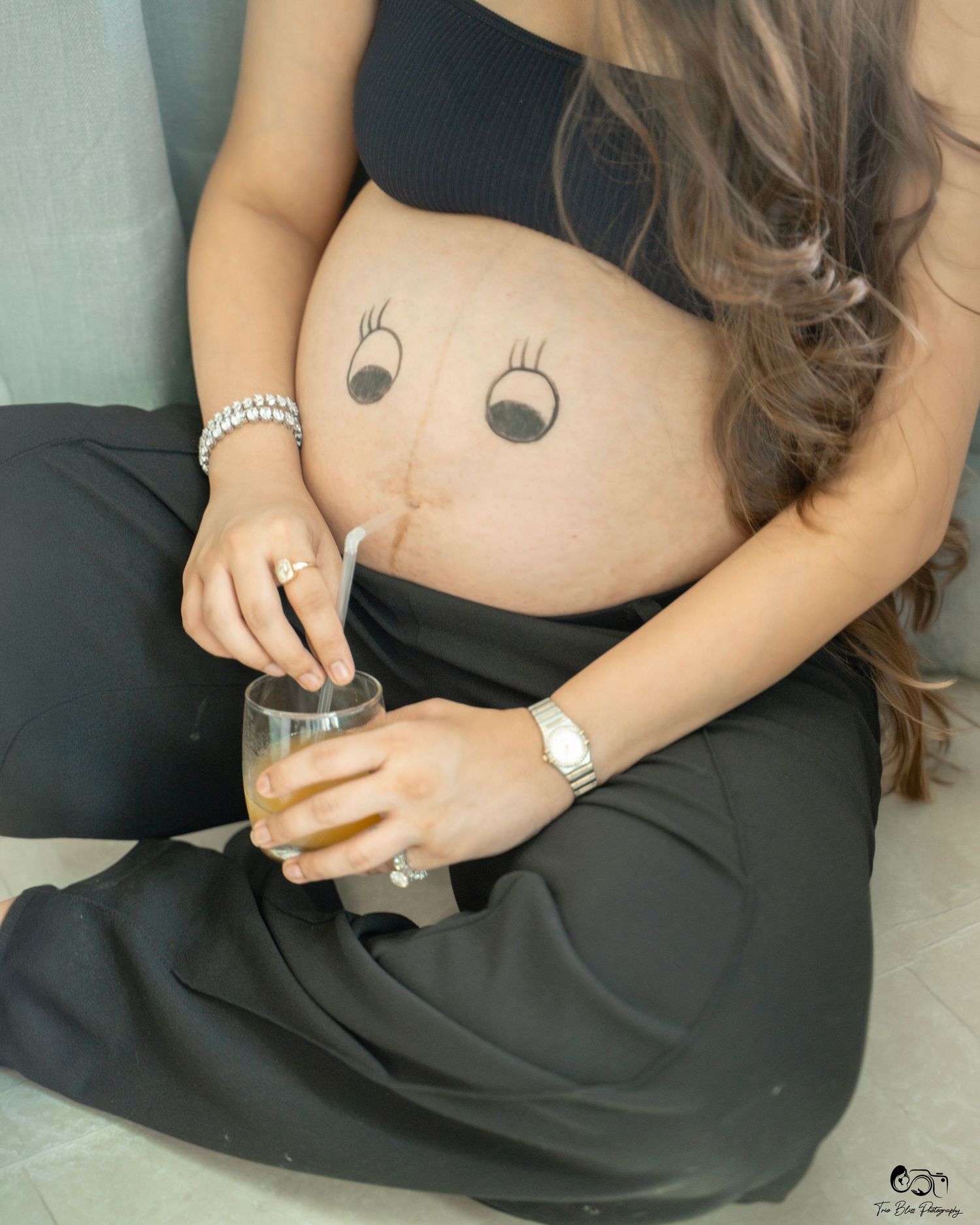 Pregnant woman in black outfit with cute eyes drawn on baby bump, holding a glass drink with a straw.