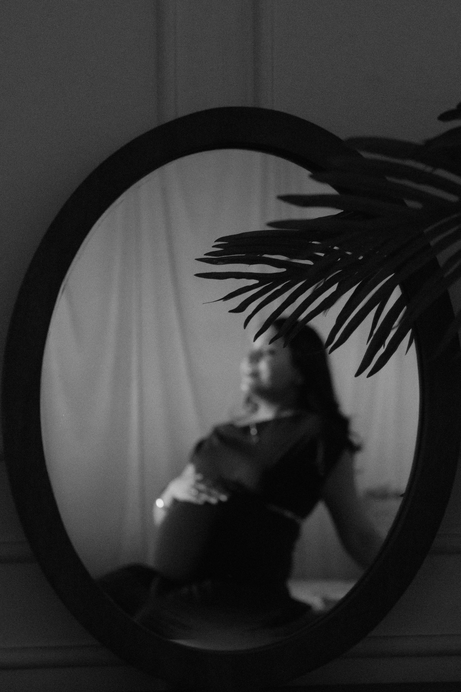 Black and white reflection of a pregnant woman posing in an oval mirror, framed by tropical palm leaves.
