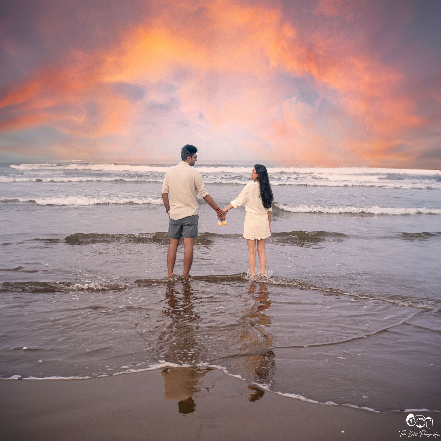 Couple holding hands on a beach at sunset with dramatic orange and pink clouds over ocean waves.