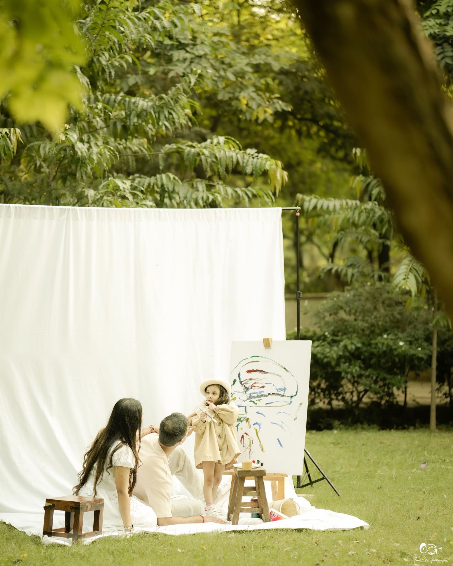 Family sitting on grass before white backdrop outdoors, with a colorful painting on easel, surrounded by lush green trees.