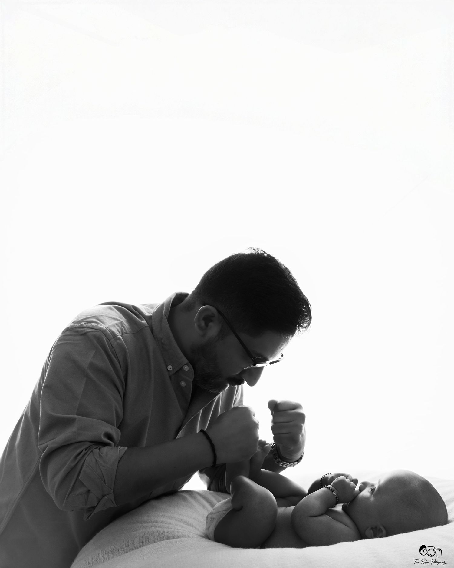 Black and white photo of a father tenderly holding his newborn baby's hands while the infant lies on a white surface.