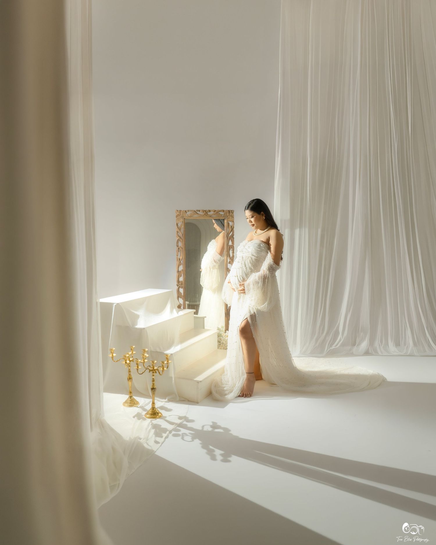 Bride in white wedding gown and fur wrap poses by ornate gold mirror with candelabra in elegant white studio.