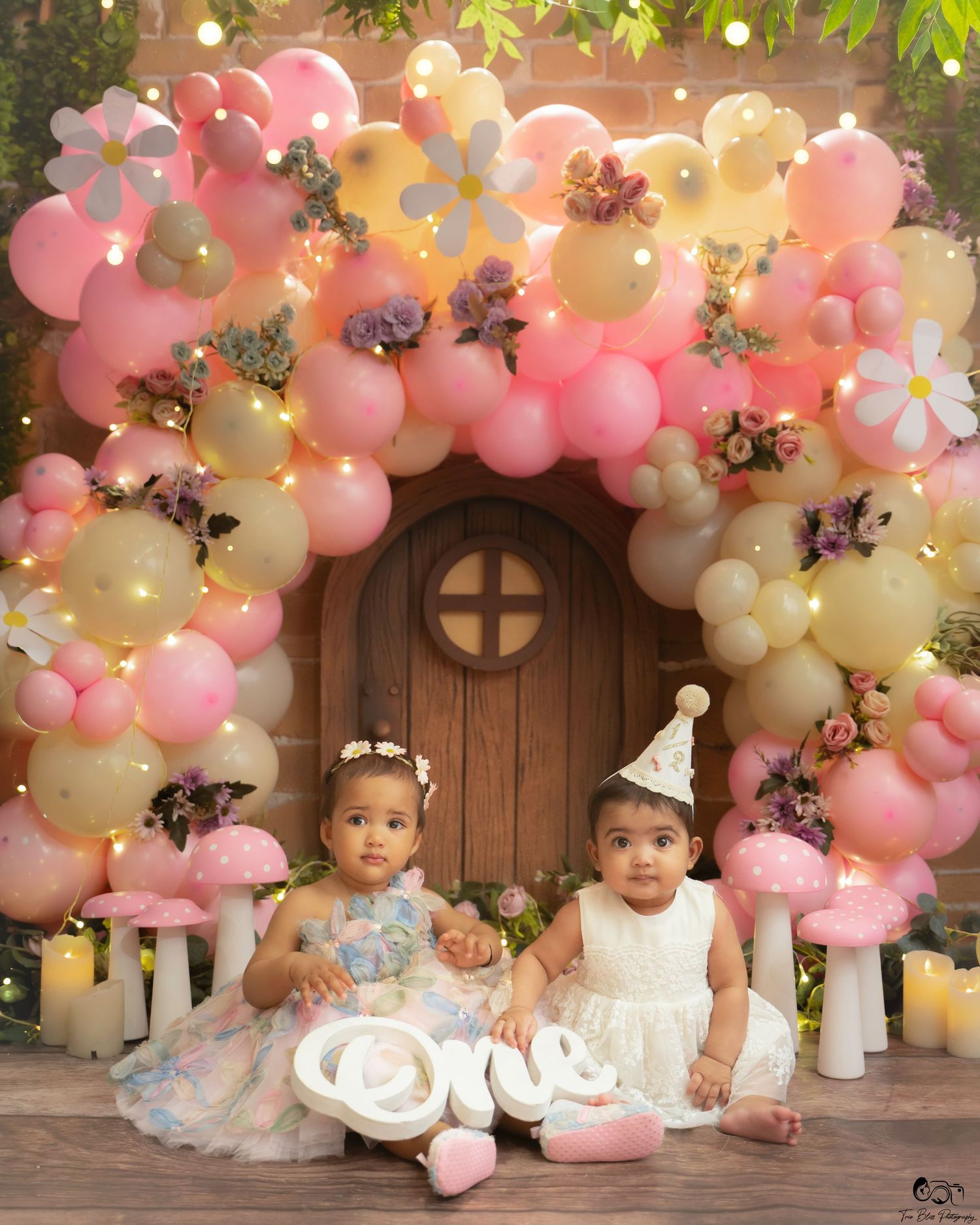 Two babies in white dresses sit under a floral balloon arch with daisy decor and an 