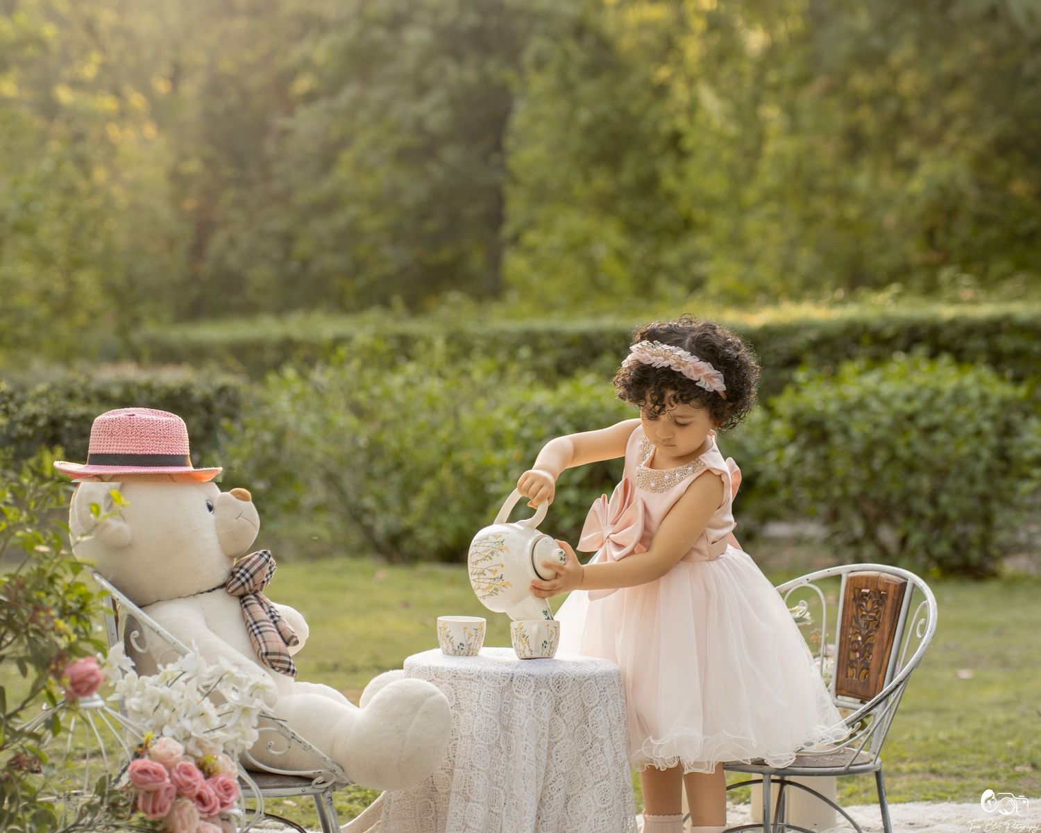 A little girl in a pink dress pours tea for a stuffed bear at an outdoor garden tea party.