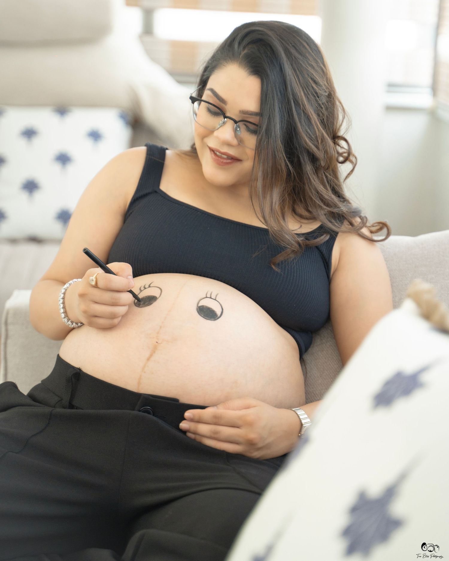 Pregnant woman drawing a cute face on her baby bump while sitting on a couch during a maternity photo session.