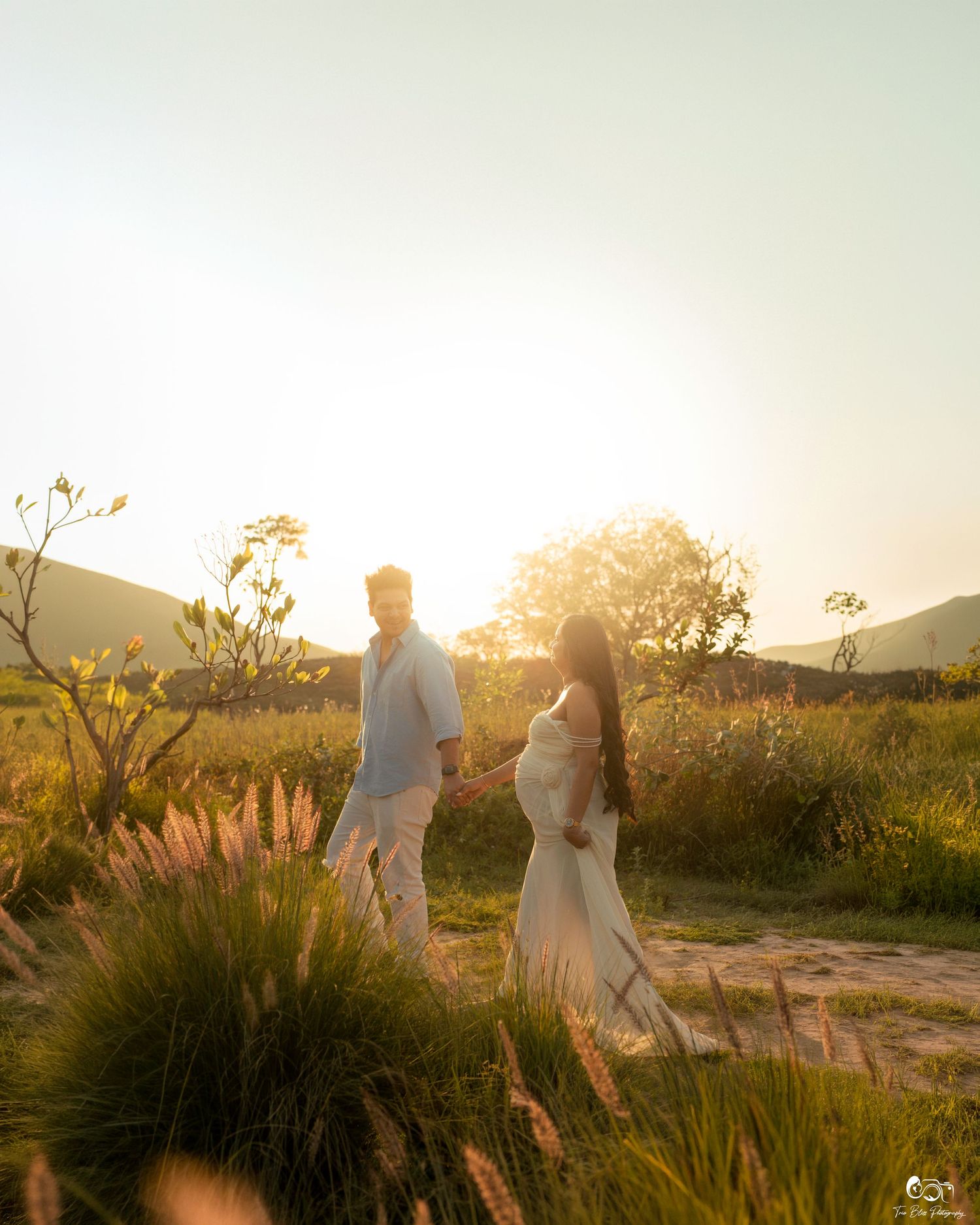 Couple holding hands in golden sunset light, pregnant woman in white dress, lush green field with mountains in background.