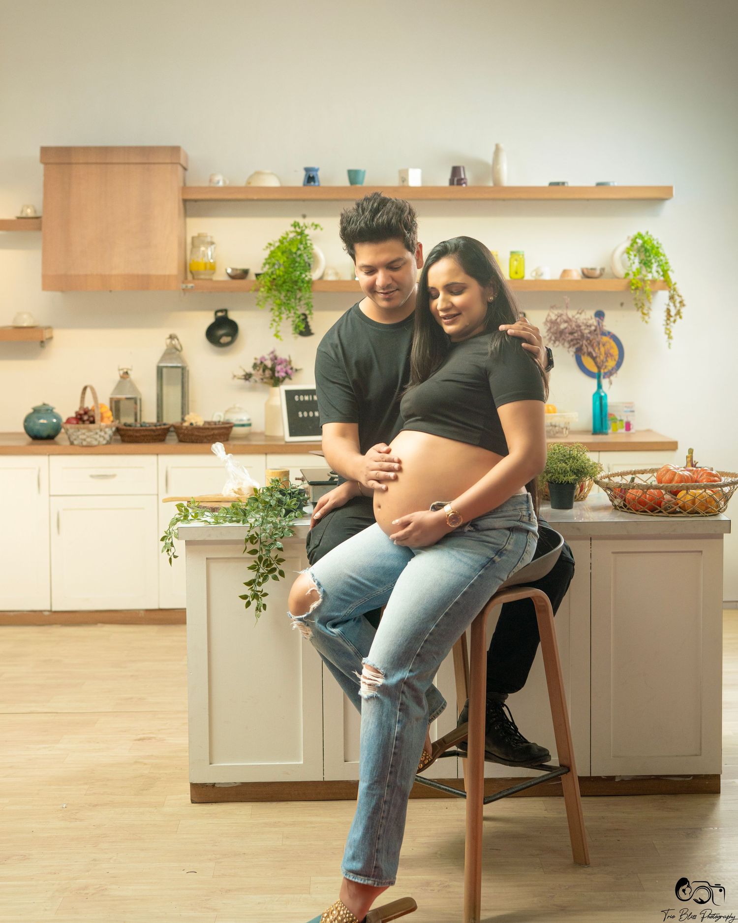 A couple shares a tender maternity photo in a modern kitchen, man embracing pregnant woman seated on a stool.