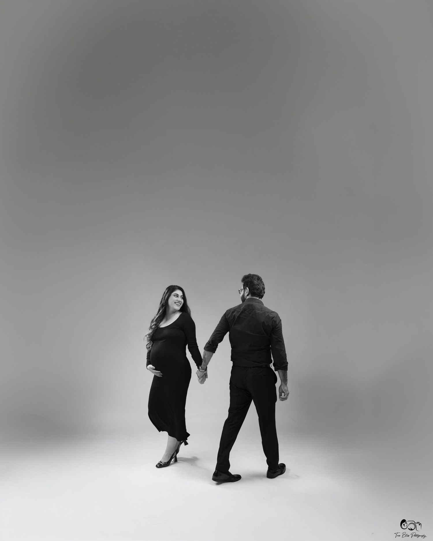 Black and white photo of a pregnant couple holding hands in a studio, woman in black dress looking back at camera.