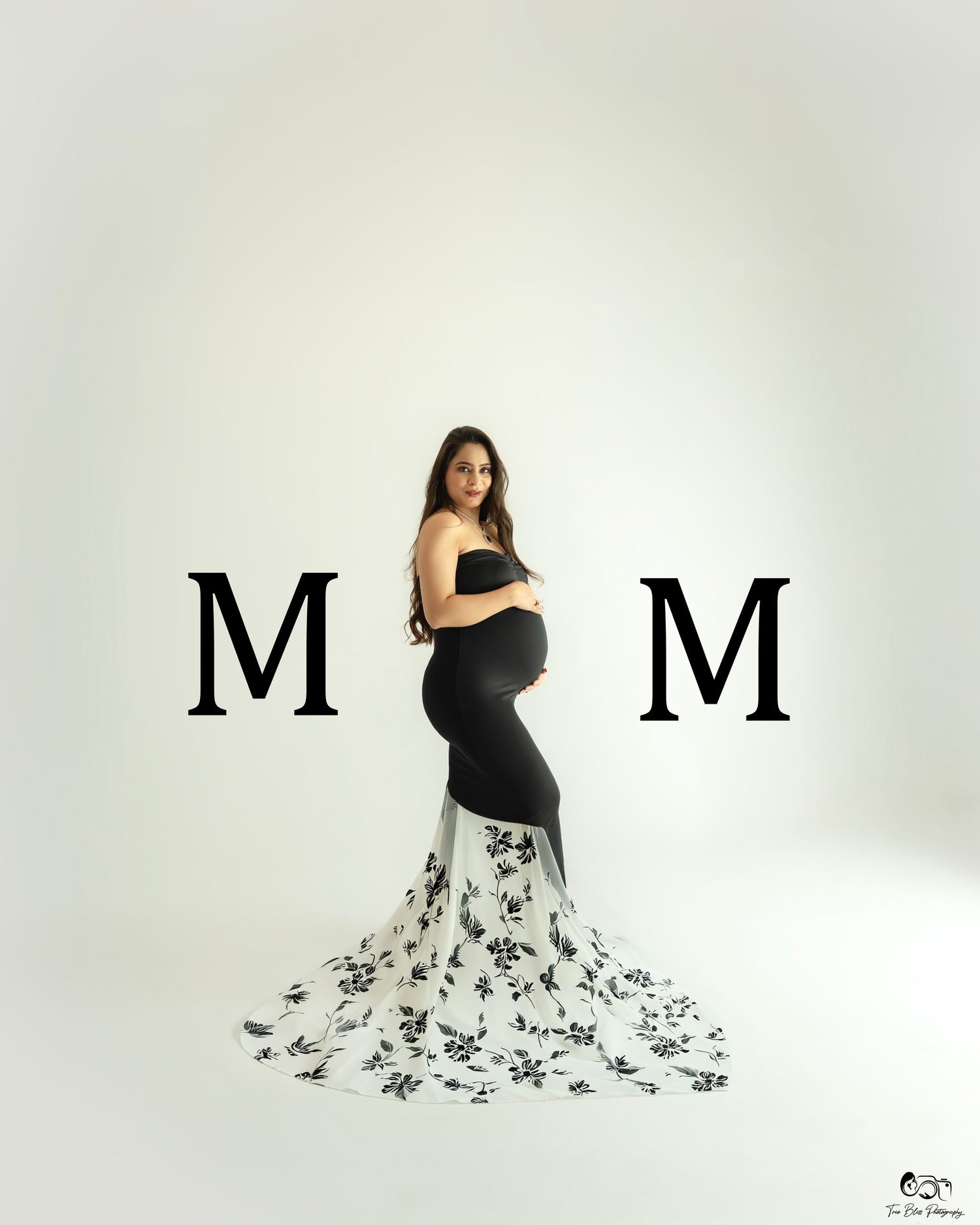 Pregnant woman in black maternity gown with floral train poses between two M letters in professional studio photo.