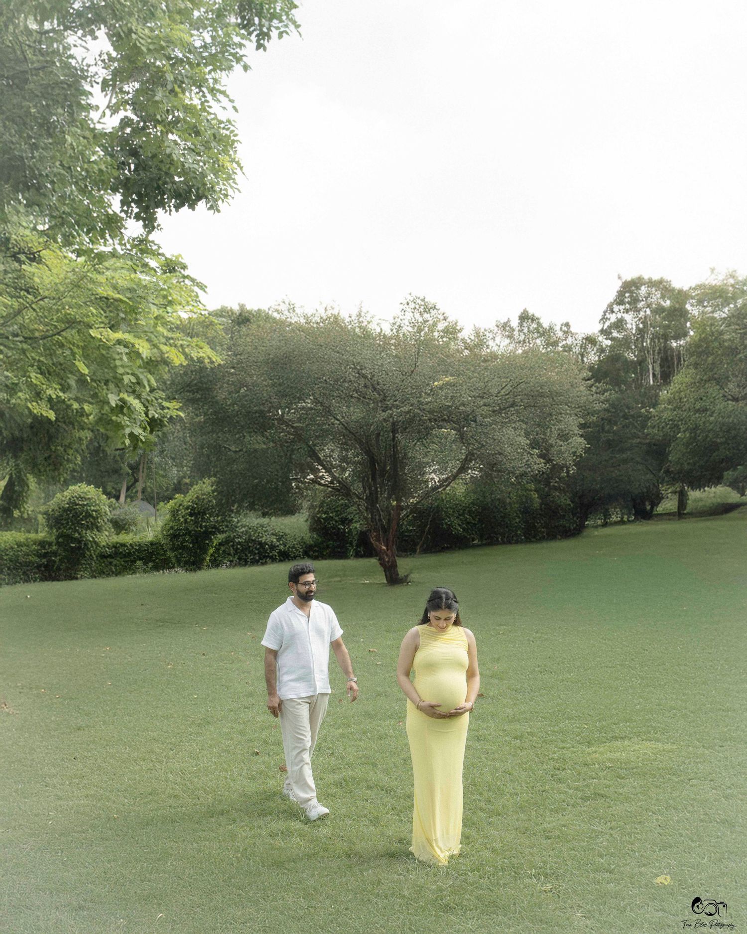A couple walks together on a lush green lawn surrounded by trees on a misty day.