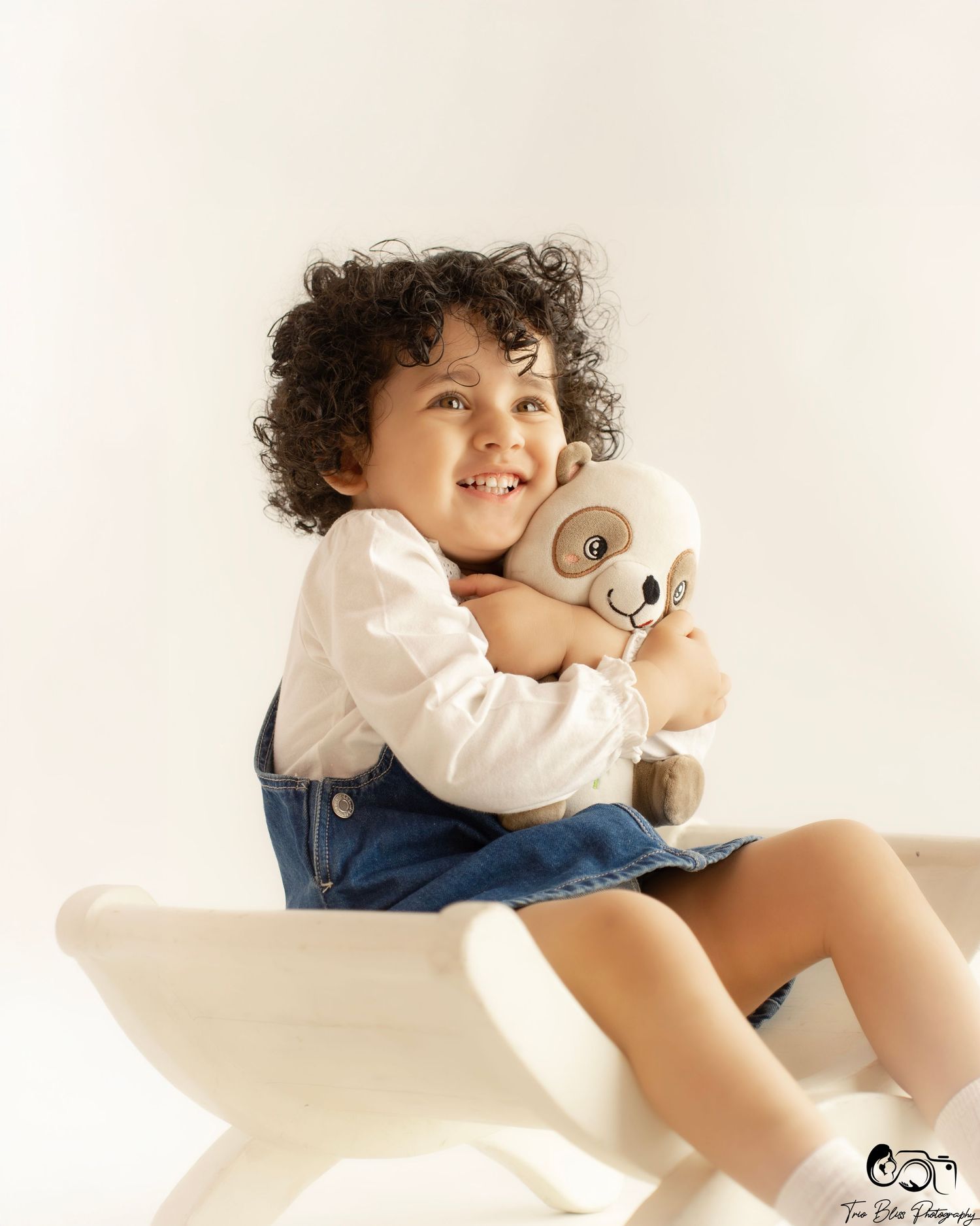 Smiling toddler with curly hair hugging a teddy bear stuffed animal while sitting on a white bench.