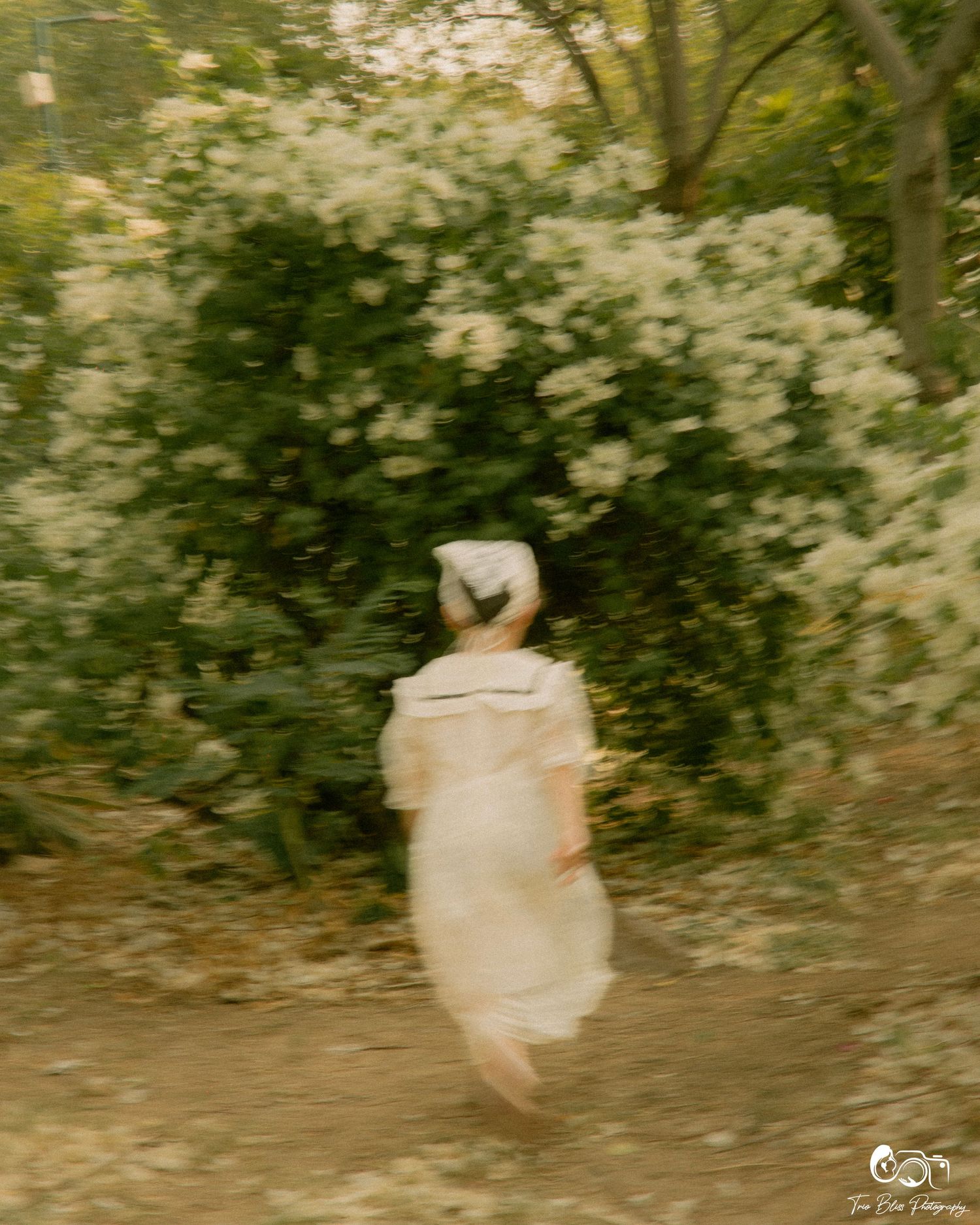 A blurred figure in a white dress runs toward blooming white flowers in a dreamy, soft-focus forest path.