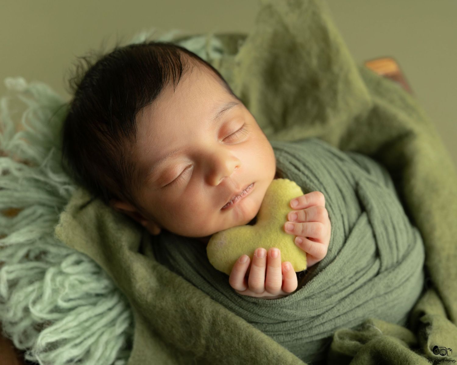 Newborn baby sleeping peacefully wrapped in green fabric, holding a small green bear toy in a cozy nest.