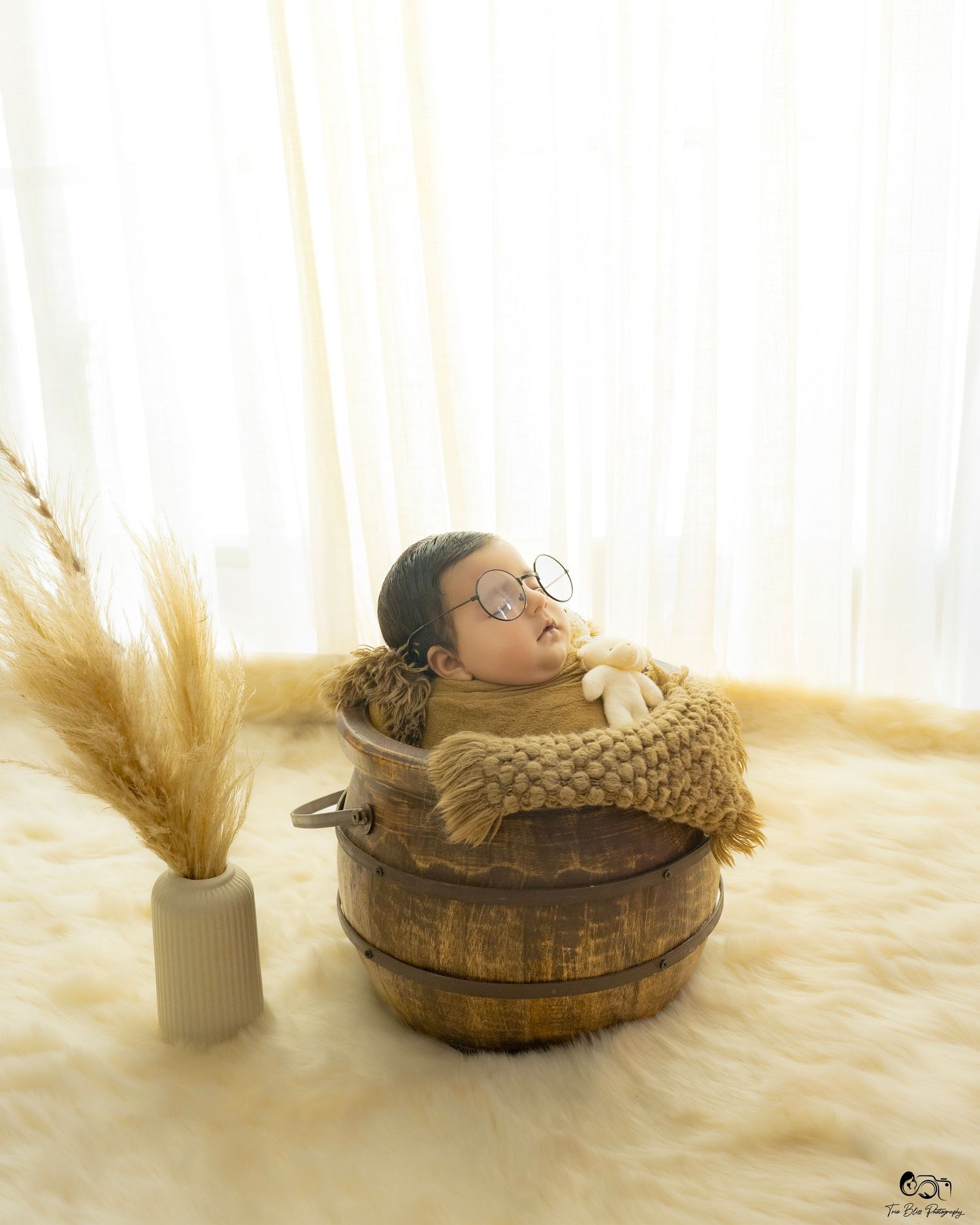 Newborn baby sleeping in a rustic wooden bucket with a knit blanket and glasses, beside dried pampas grass on a fluffy rug.