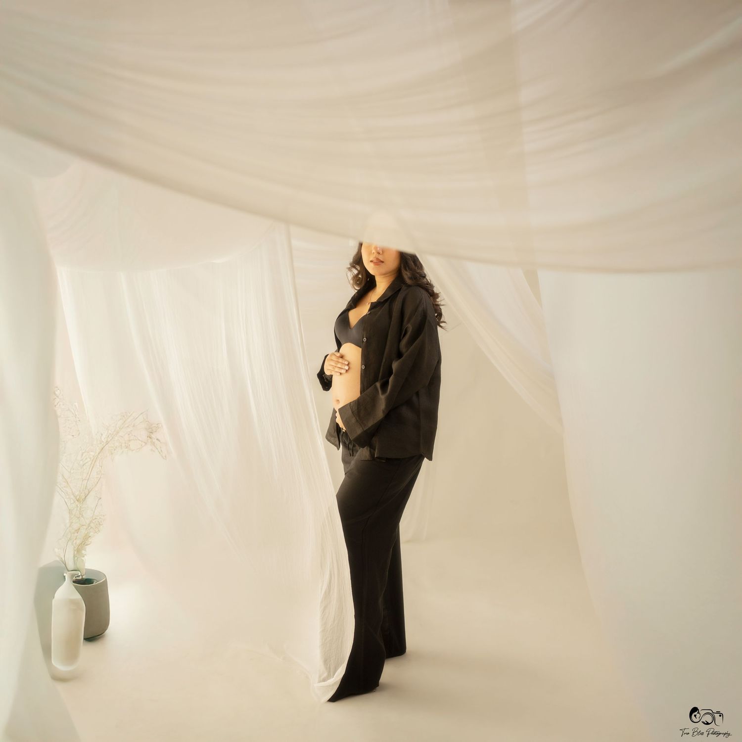 A person wearing black clothing stands in a dreamy white fabric-draped setting with a white vase in the background.