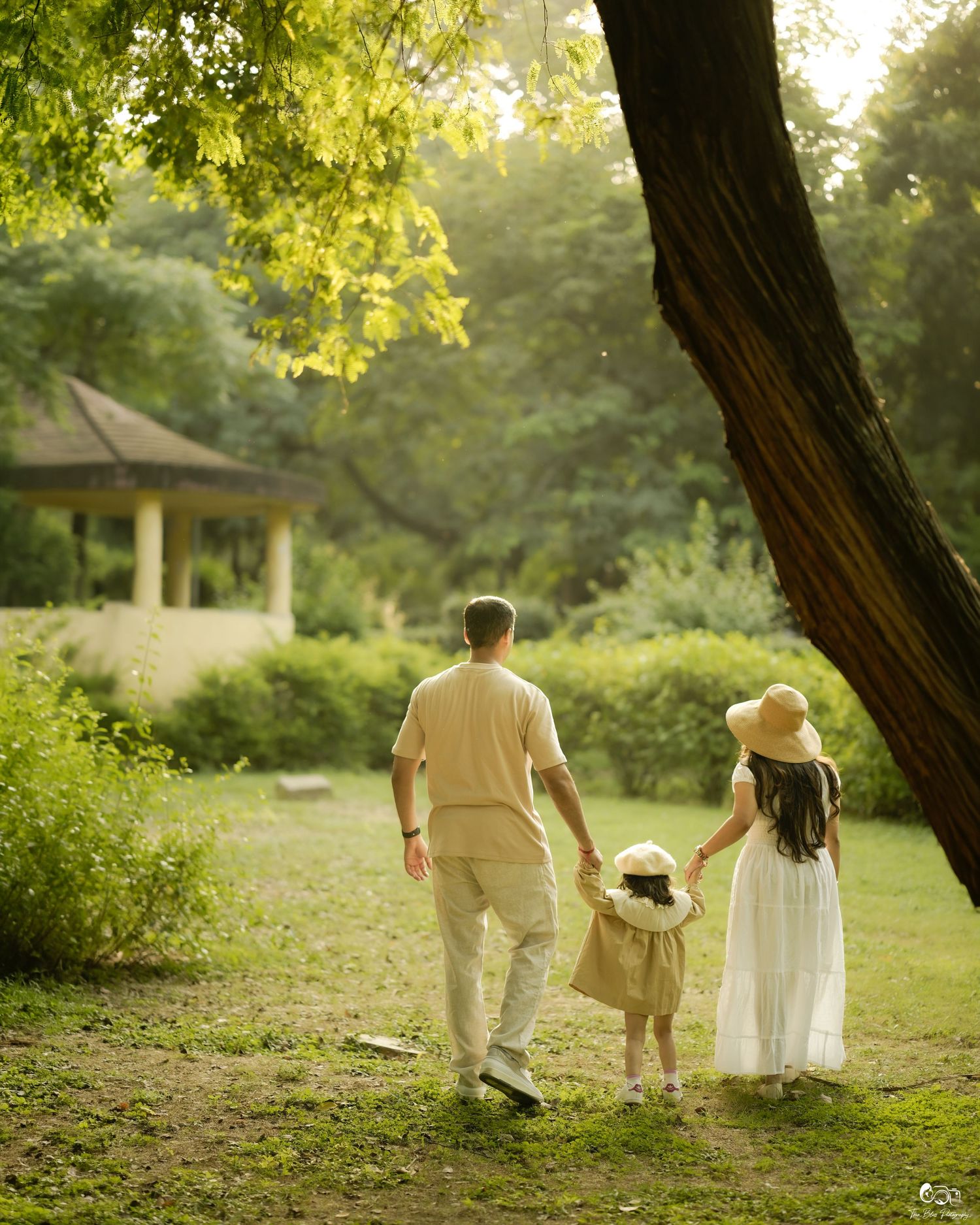 A family of three walks hand-in-hand through a lush green park, with a gazebo visible in the background.