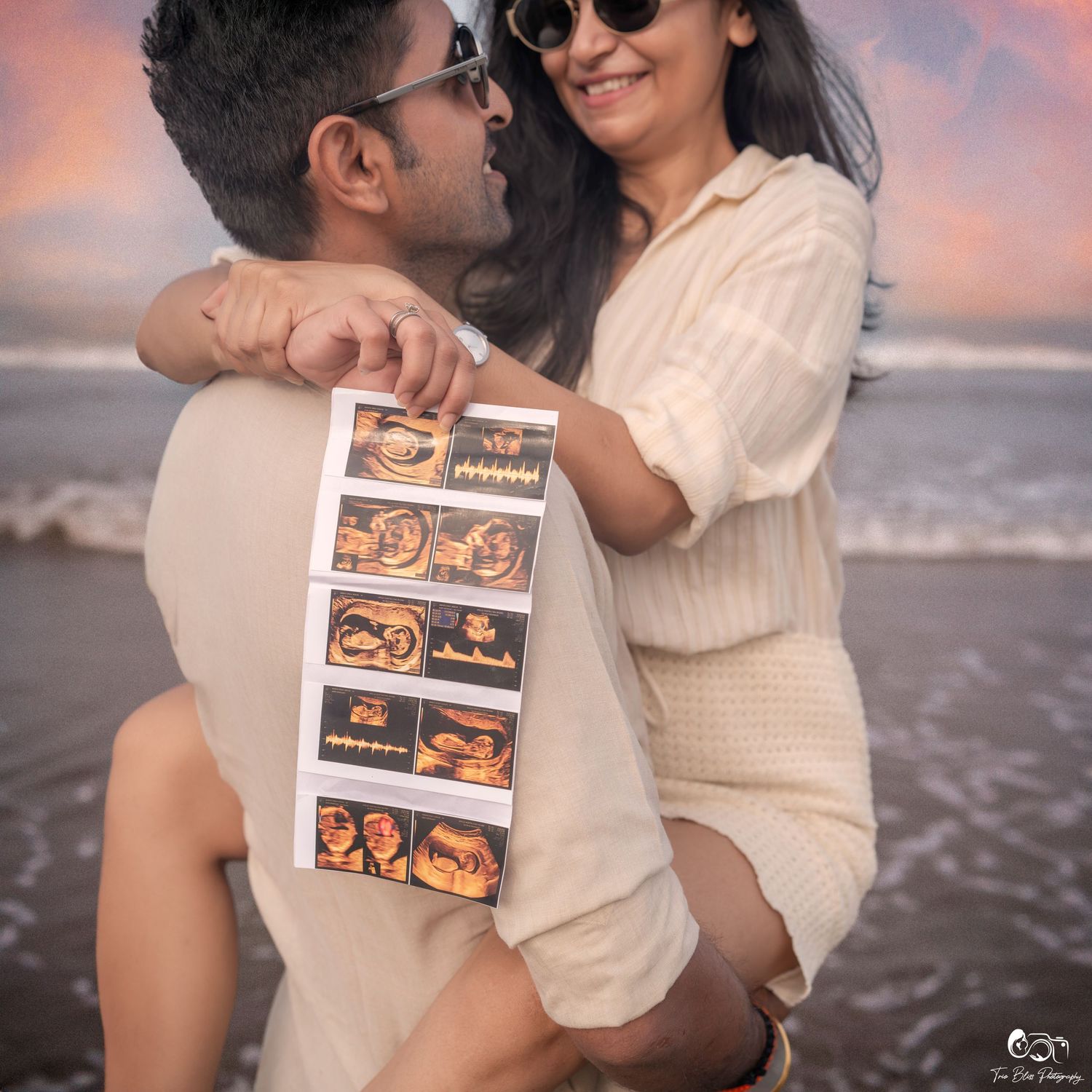 Couple on beach holding ultrasound photos, announcing pregnancy, with ocean waves and colorful sunset sky behind them.
