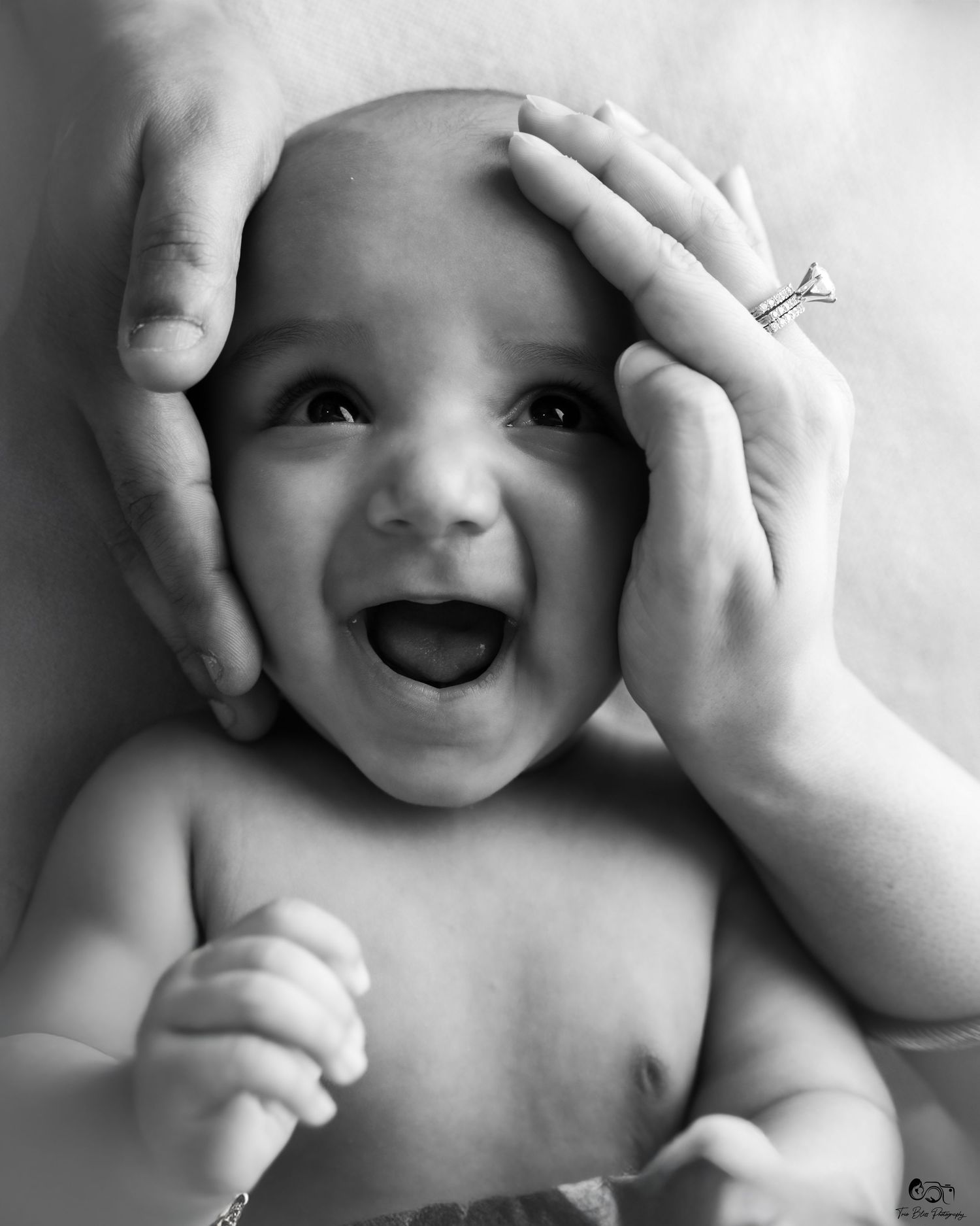 Black and white photo of a joyful laughing baby with adult hands gently cradling its face, one hand wearing a diamond ring.