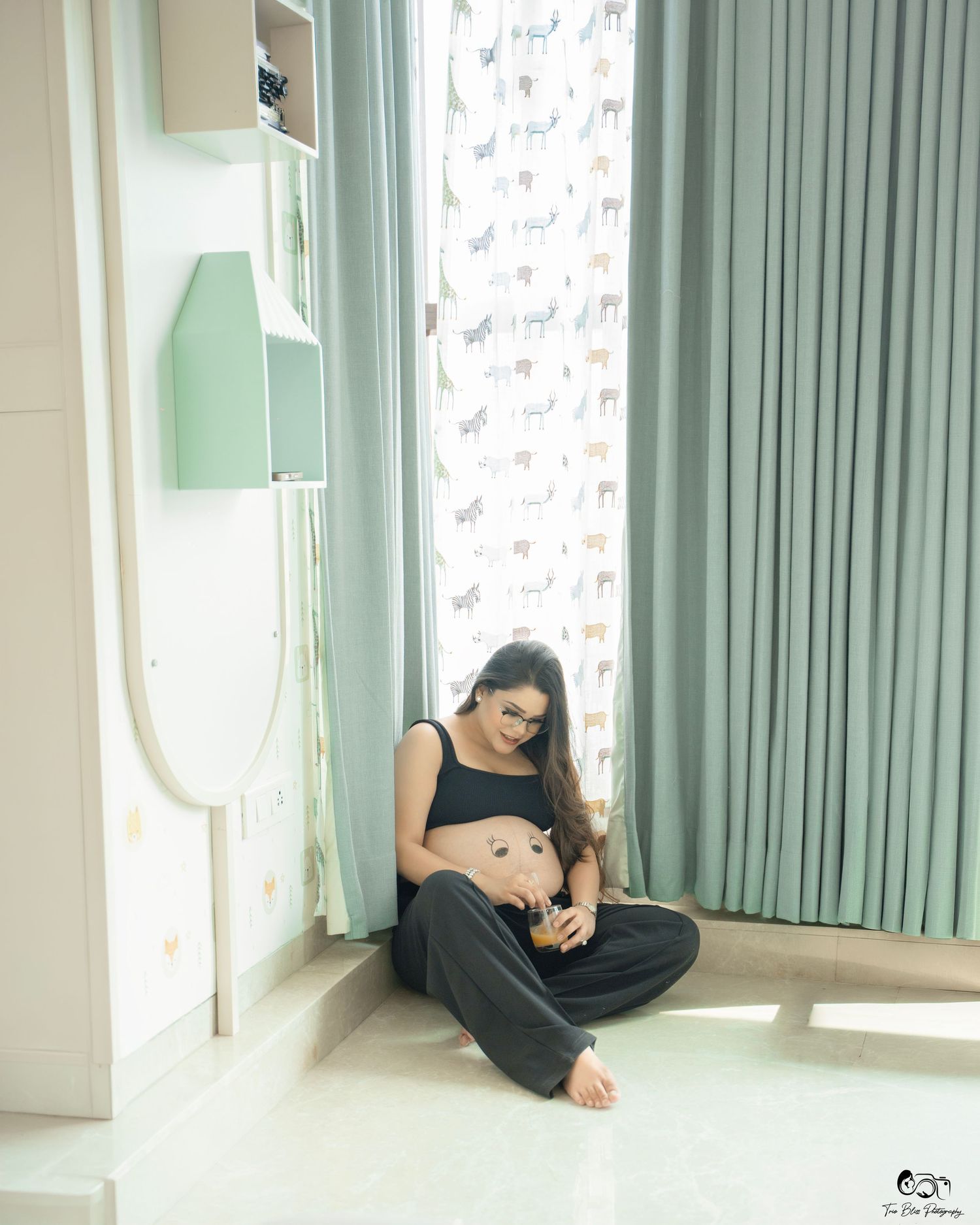 Pregnant woman sitting cross-legged on floor, cradling baby bump, wearing black crop top and pants near green curtains.