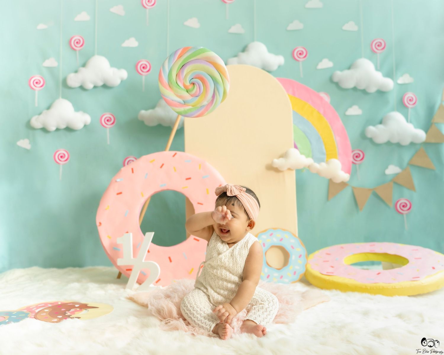 Baby girl sits among giant donut props and lollipops in a colorful candy-themed photo shoot with rainbow backdrop.