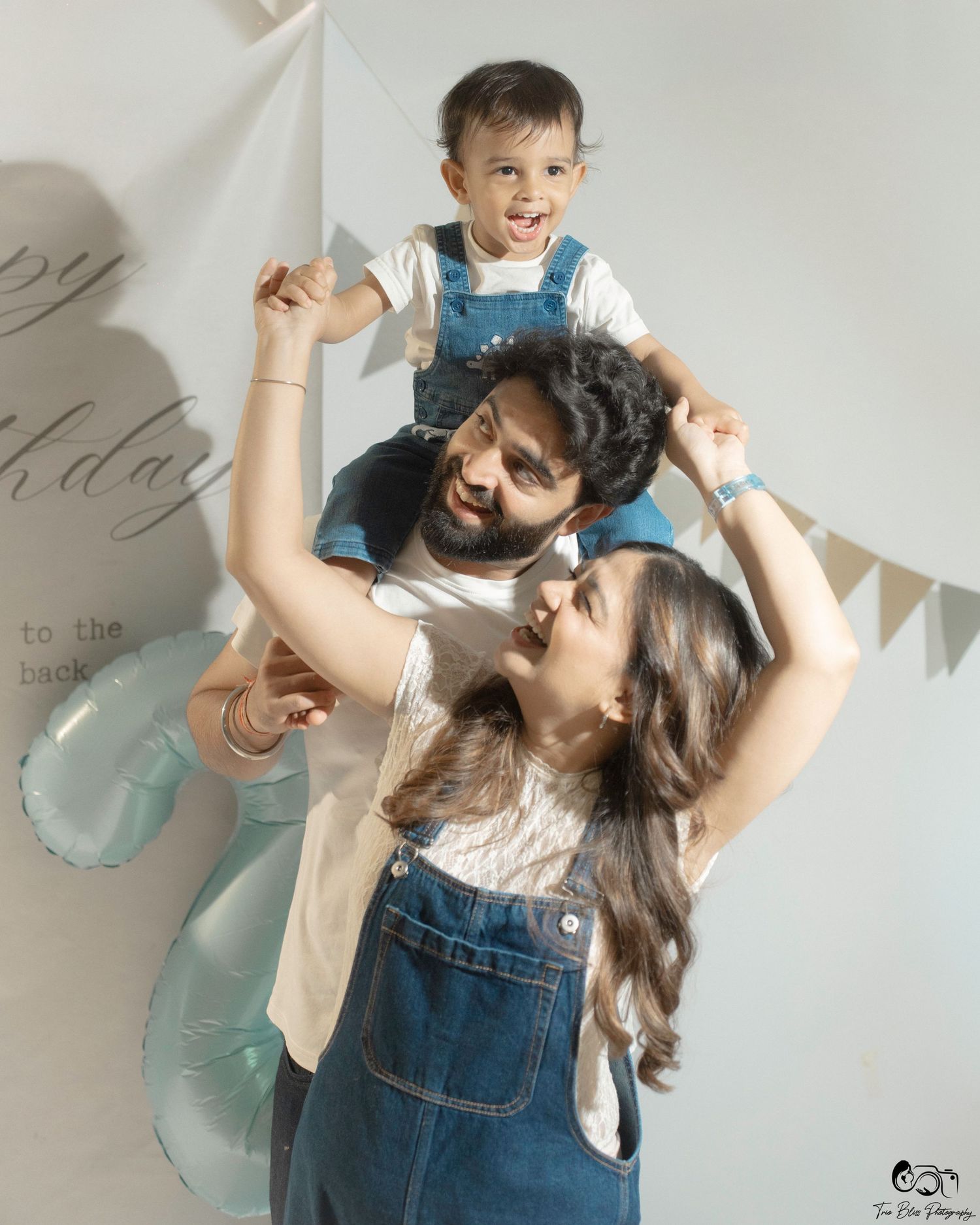 A joyful family photo with parents in denim overalls lifting their toddler at a birthday celebration.