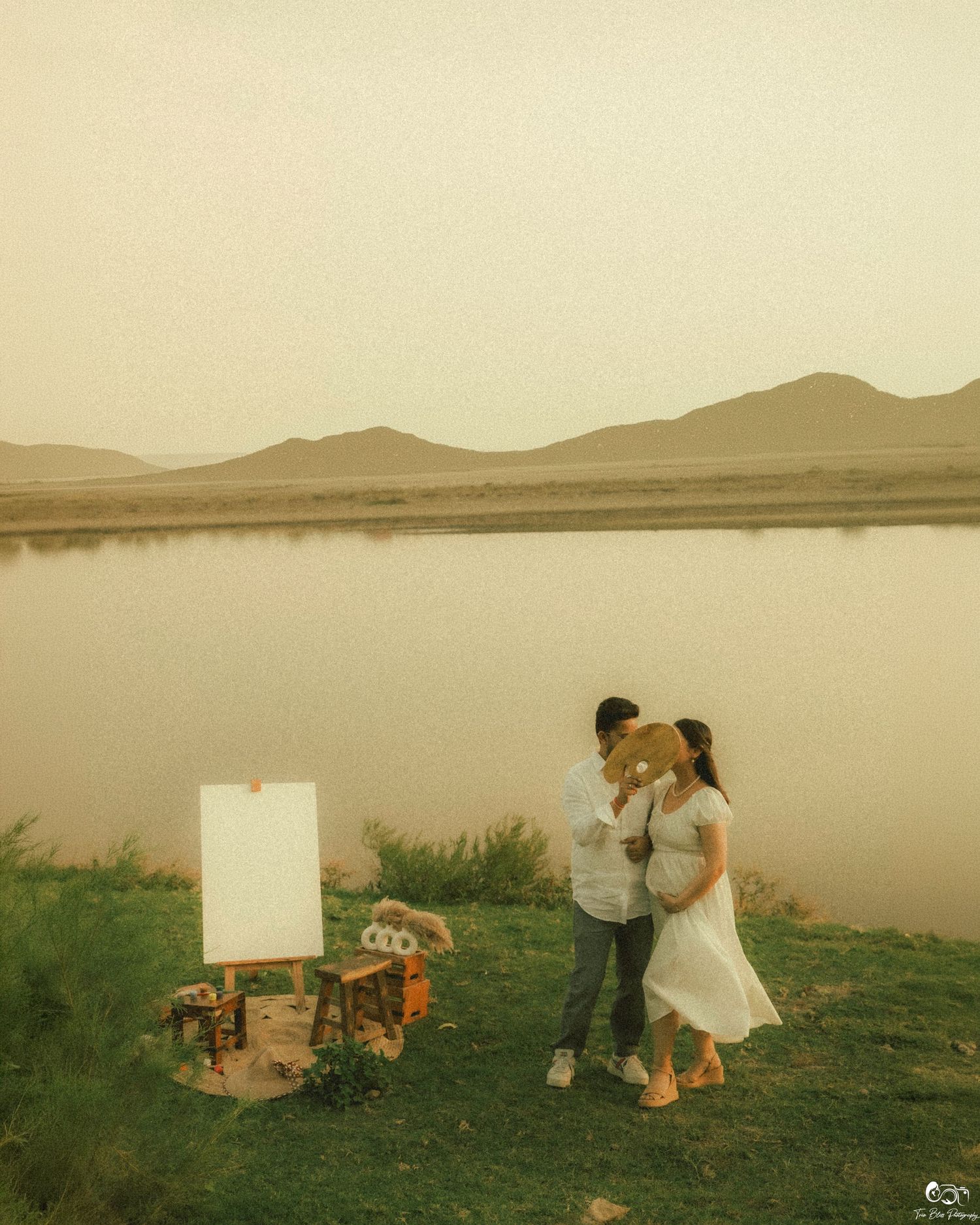 Pregnant couple sharing a tender moment by a serene lake with mountains, beside a blank canvas easel at golden hour.