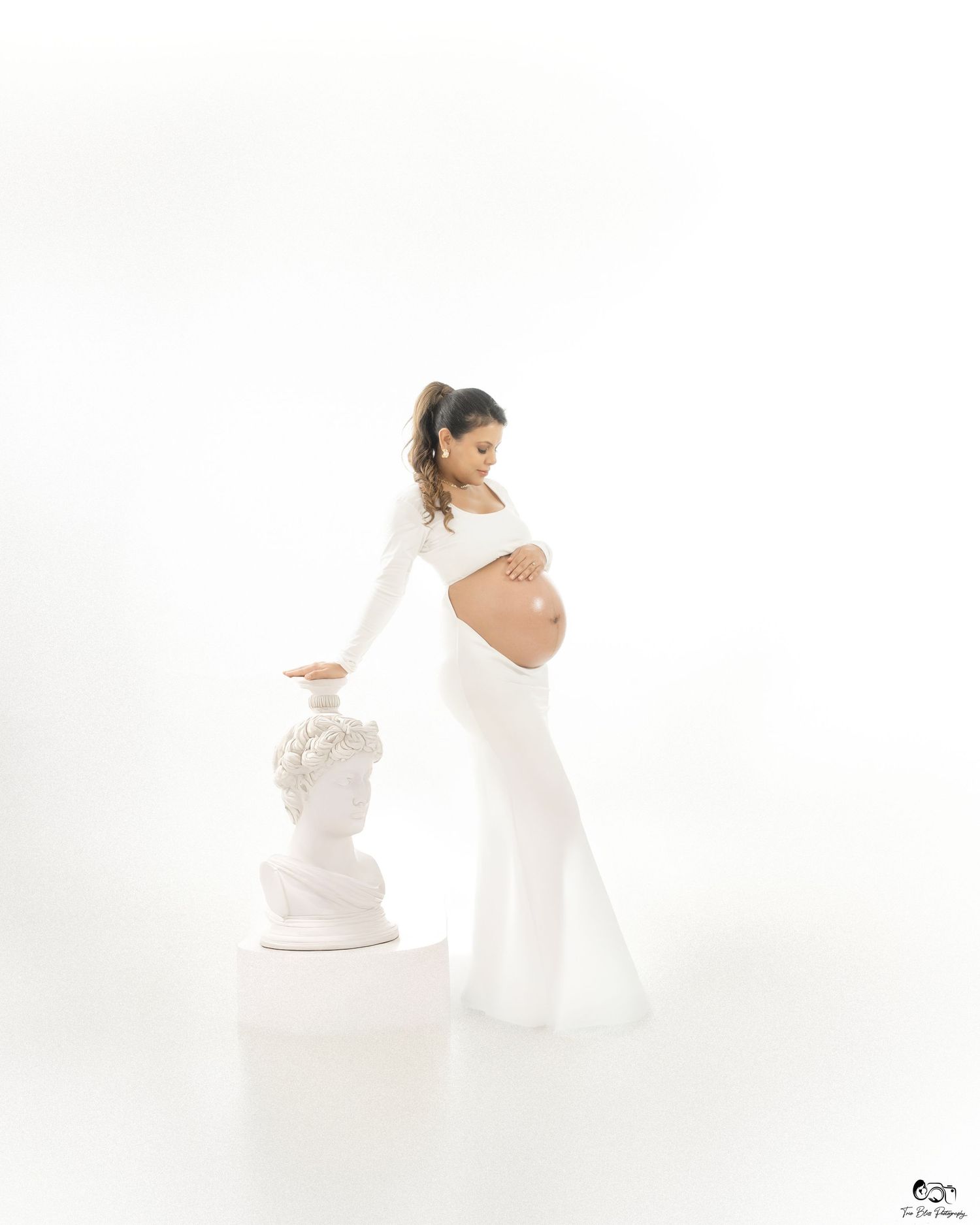 Pregnant woman in white crop top and flared pants poses gracefully beside a classical bust sculpture in a bright studio.