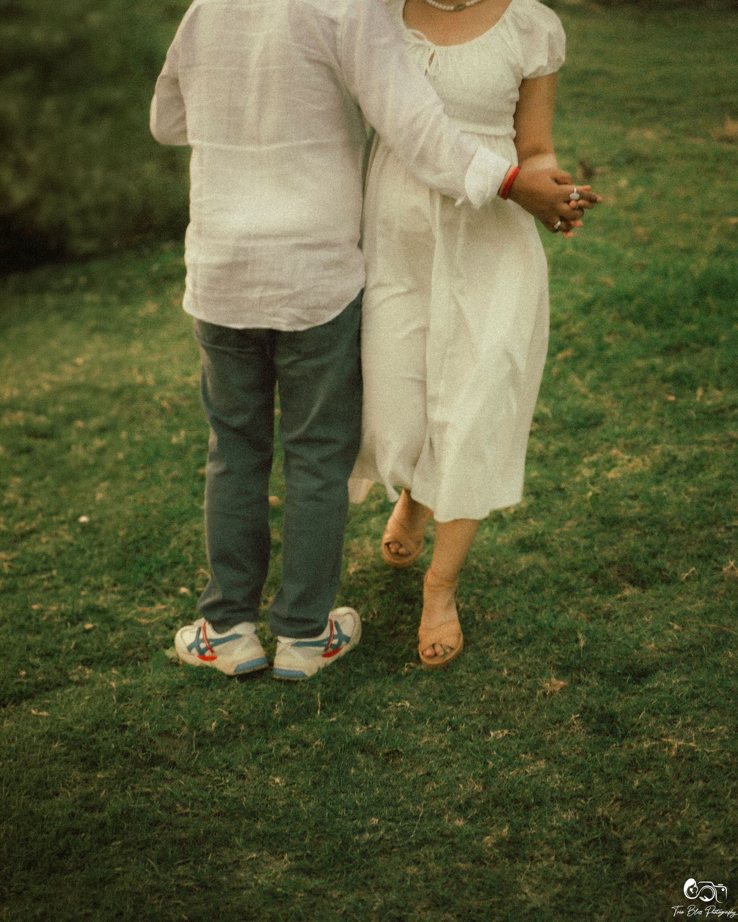 Two children in white outfits embrace on green grass, one wearing colorful sneakers and the other in sandals.