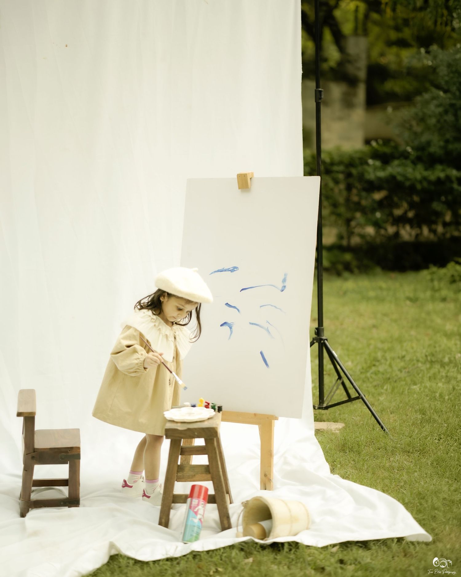 Young girl in beige coat and beret painting blue strokes on canvas easel outdoors on white blanket.