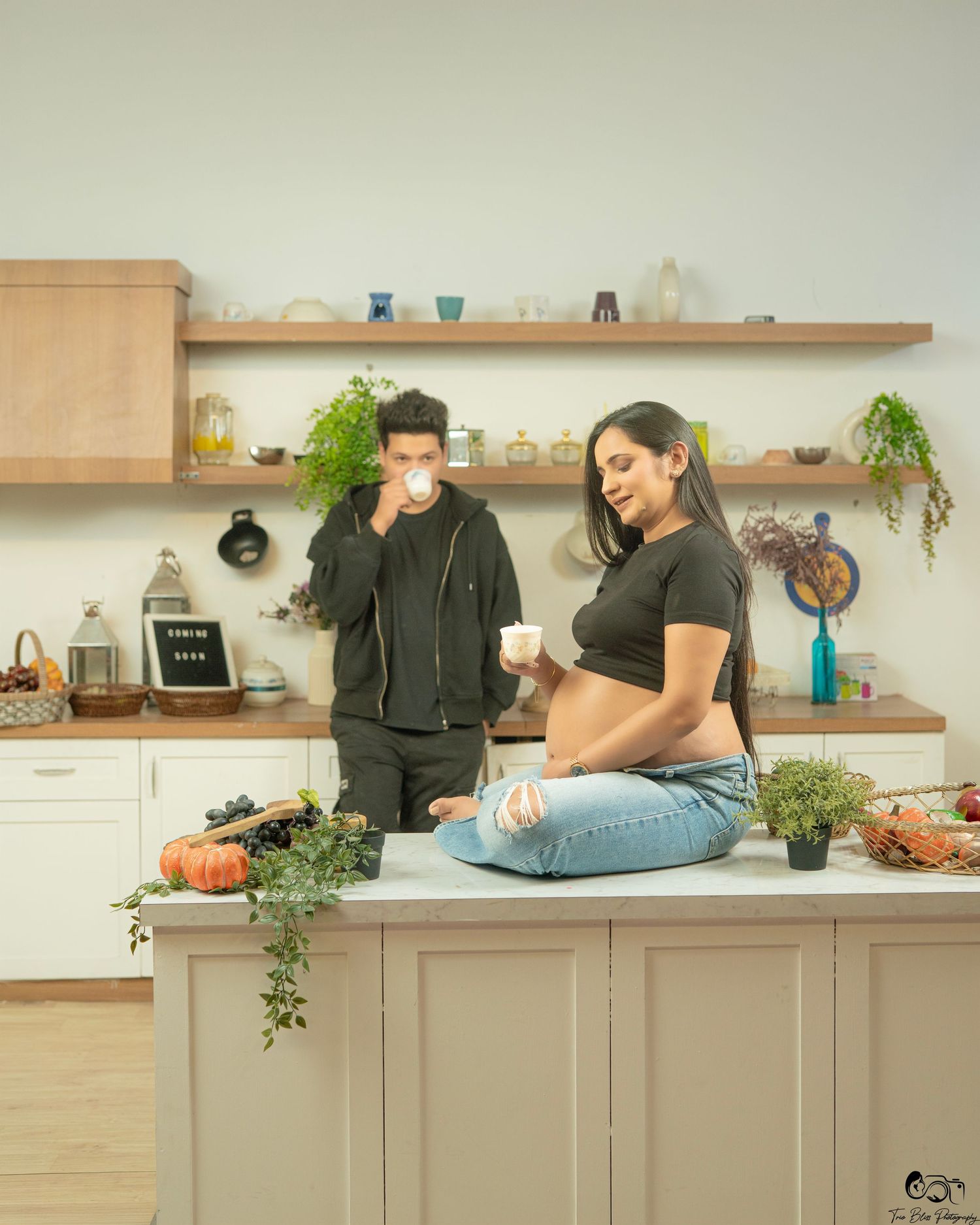 Couple in a modern kitchen, pregnant woman sitting on counter holding a cup while man drinks coffee beside her.