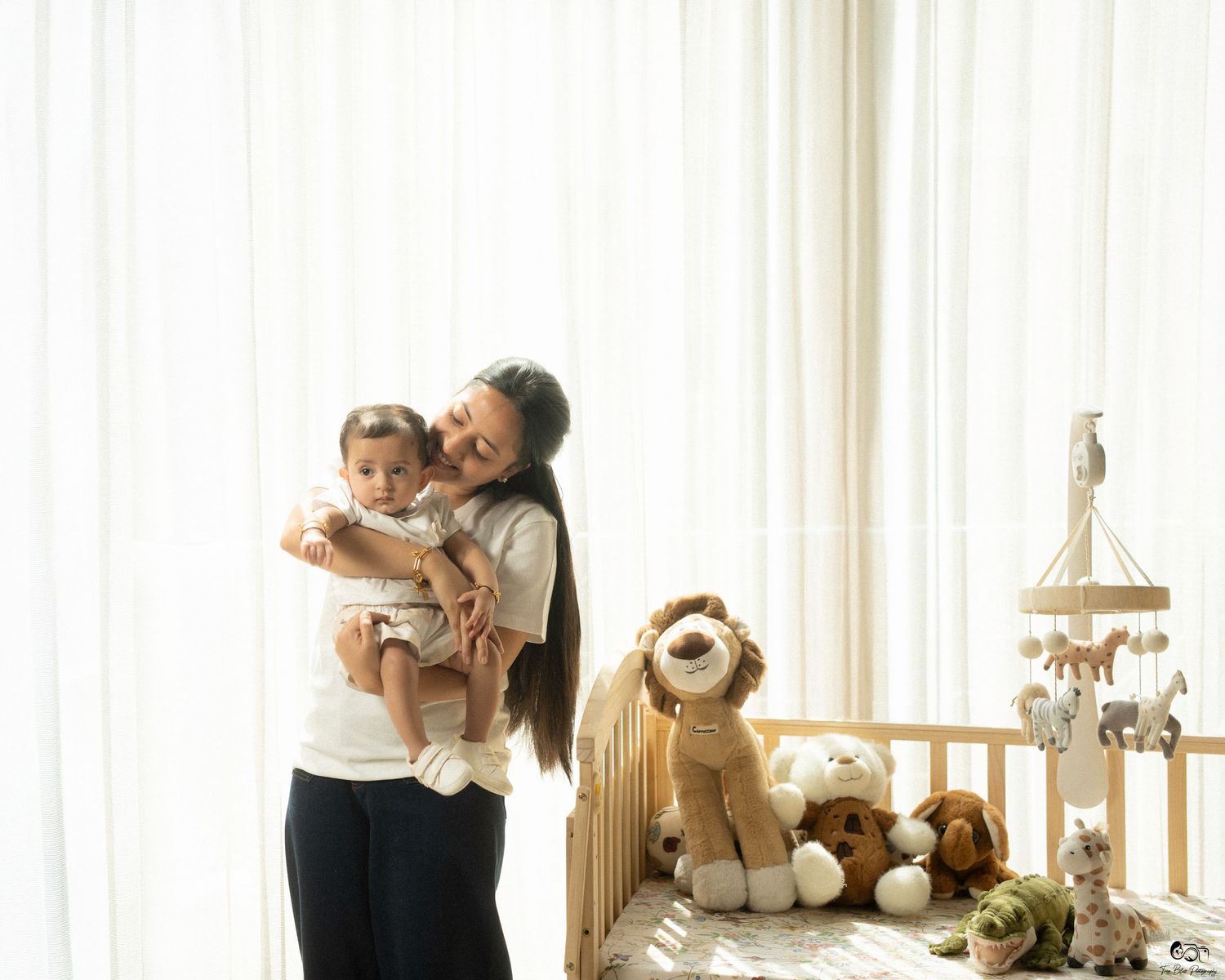 Mother holding baby girl near a wooden crib with stuffed animals by sheer white curtains in a nursery.