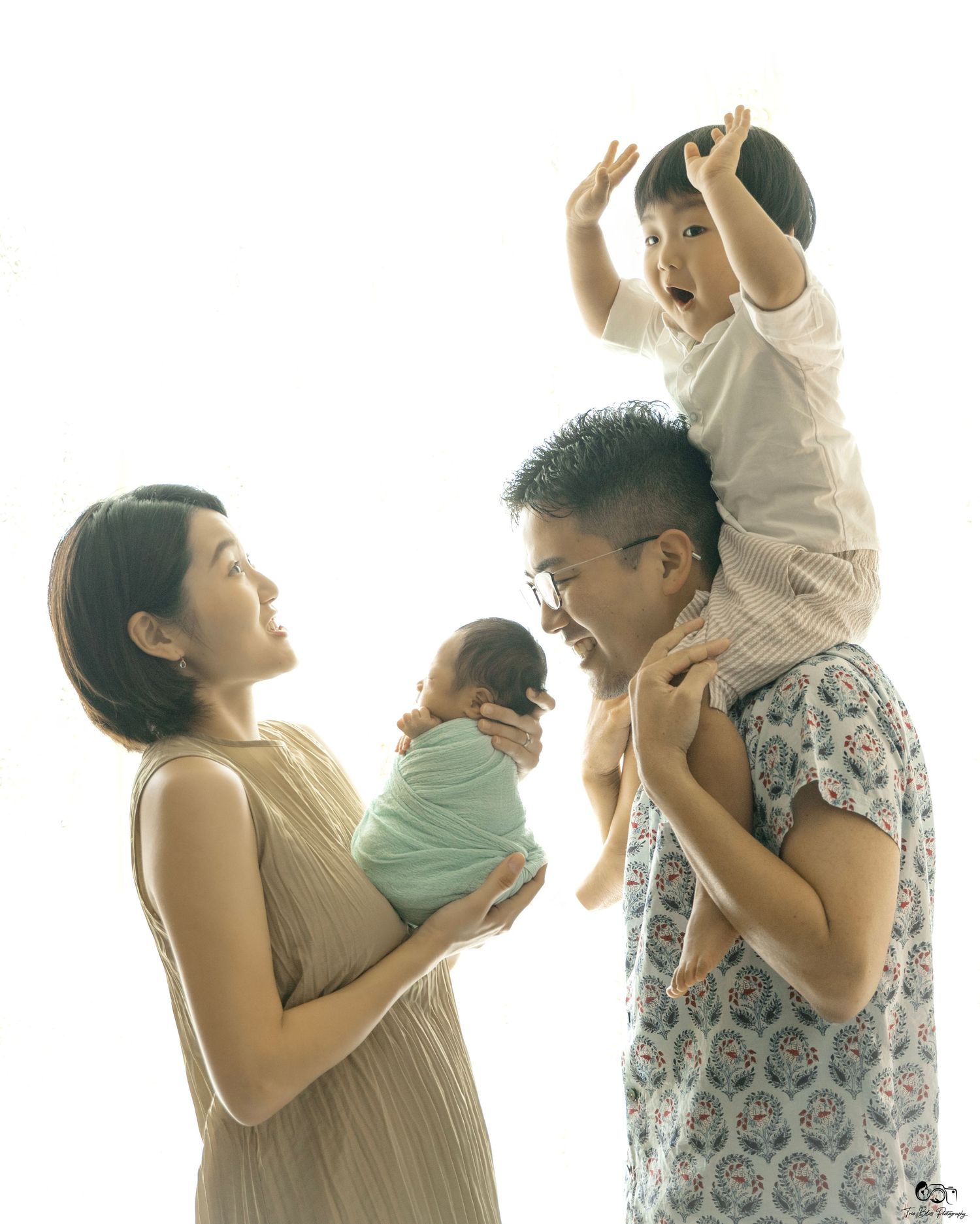 Family portrait with mother holding newborn, father with toddler on shoulders, bright white background.
