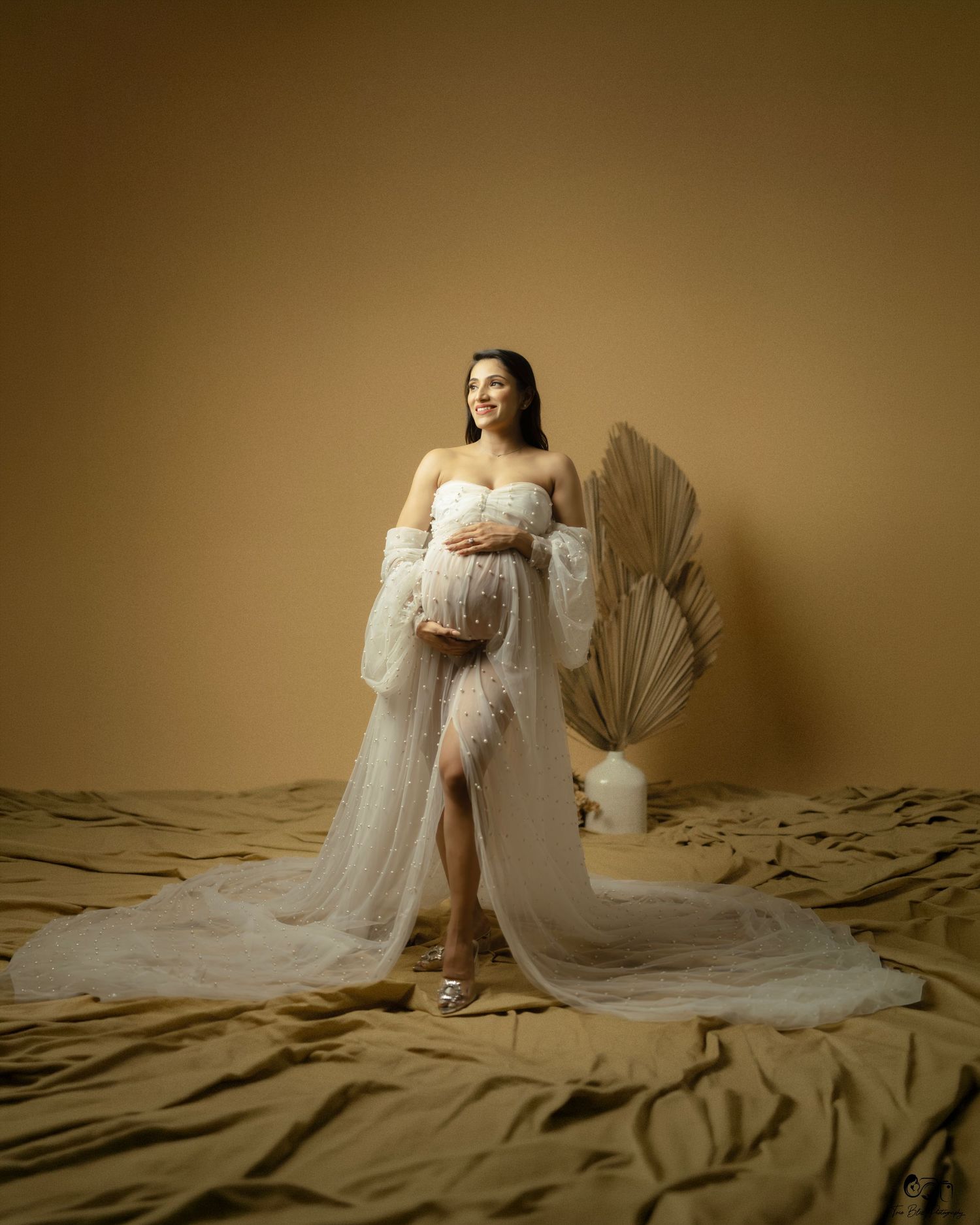Pregnant woman in white tulle maternity gown cradling baby bump in golden studio setting with pampas grass decor.